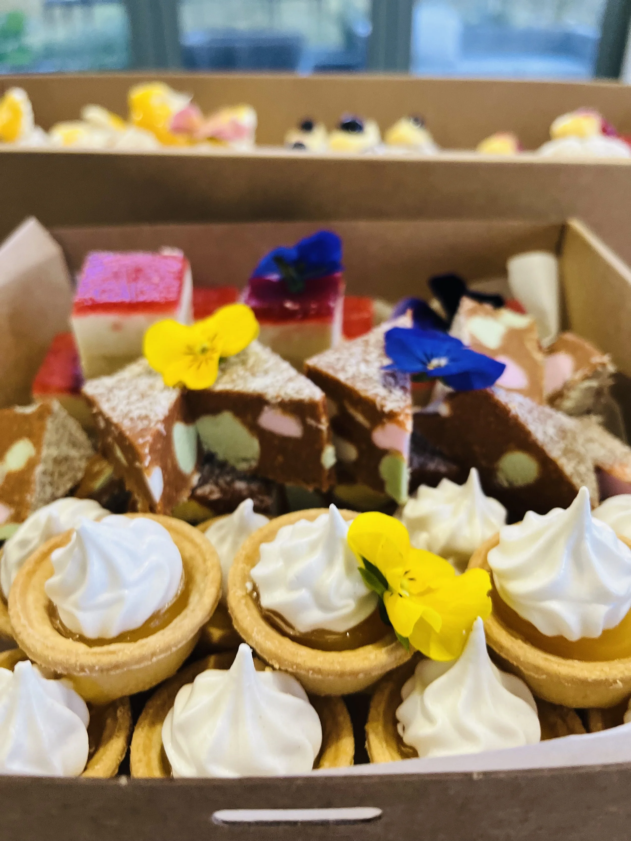 A box of assorted desserts including tartlets with white icing and edible flowers, mini slices of layered cake, and small decorated pastries.