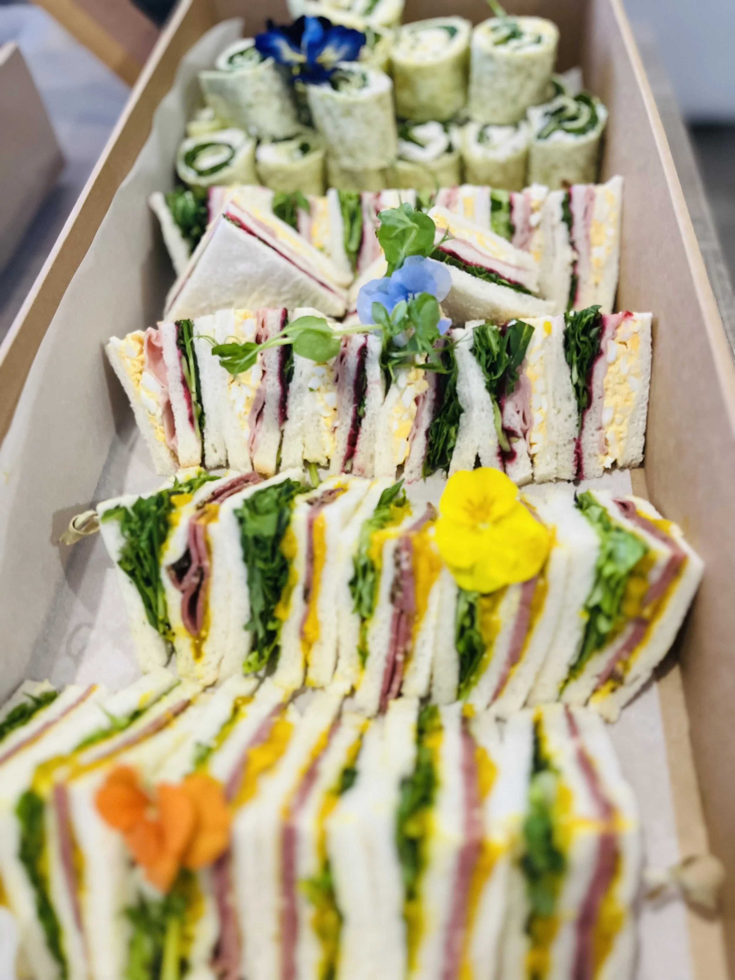 Assorted finger sandwiches arranged in a box, garnished with edible flowers and green leaves.