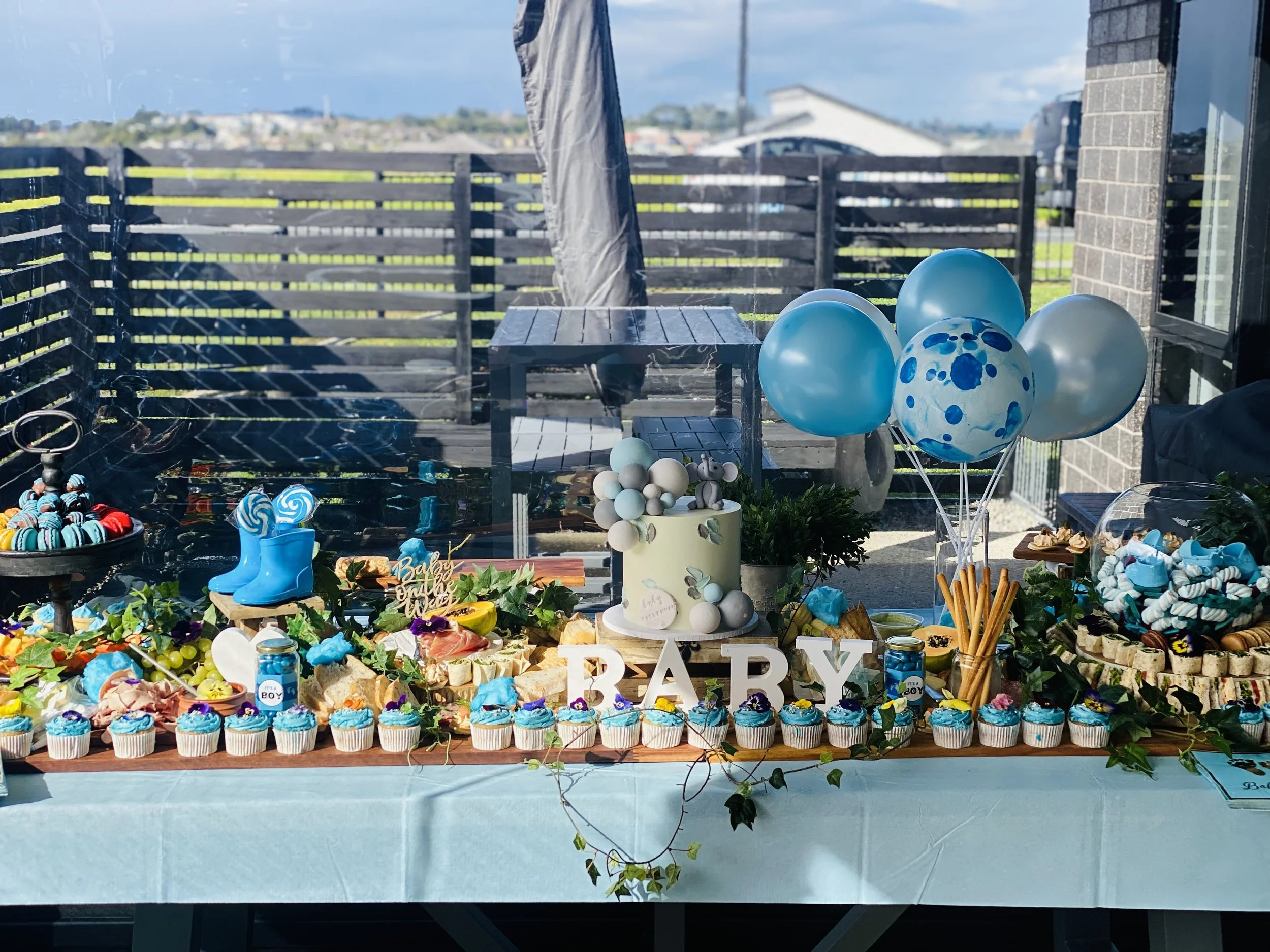 Baby shower dessert table with cupcakes, balloons, and decorations, featuring a small cake, blue and white color theme, and a sign that says 'Baby'.