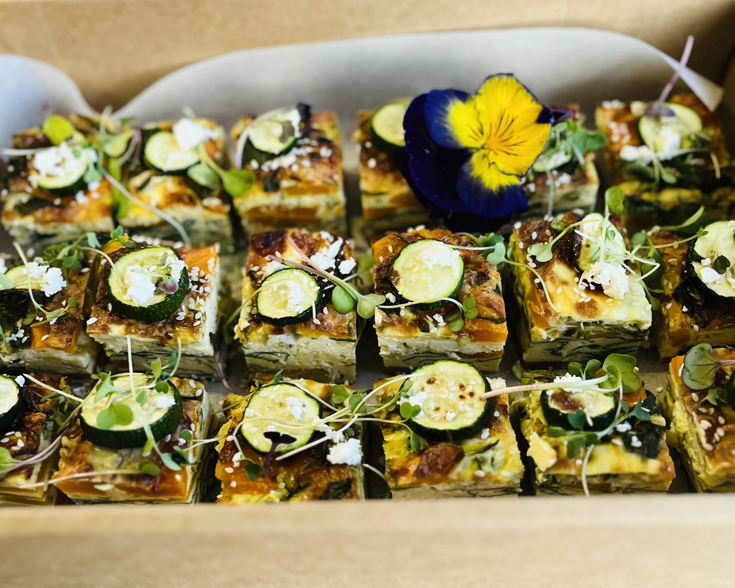 Tray of assorted savory layered appetizer squares garnished with cucumber slices, microgreens, and an edible flower.