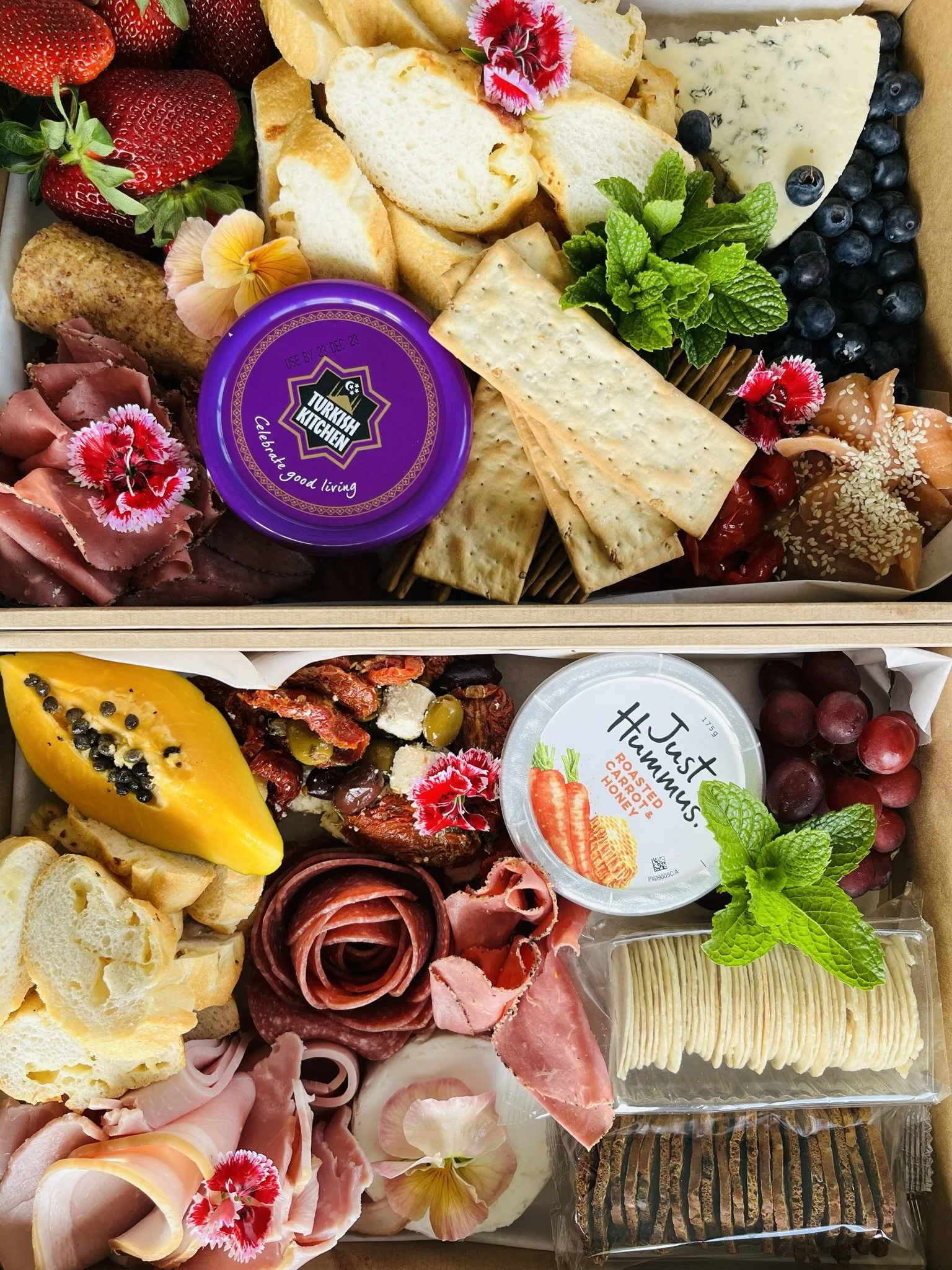Two boxes of assorted charcuterie with cheeses, fruits, crackers, meats, and garnishes.