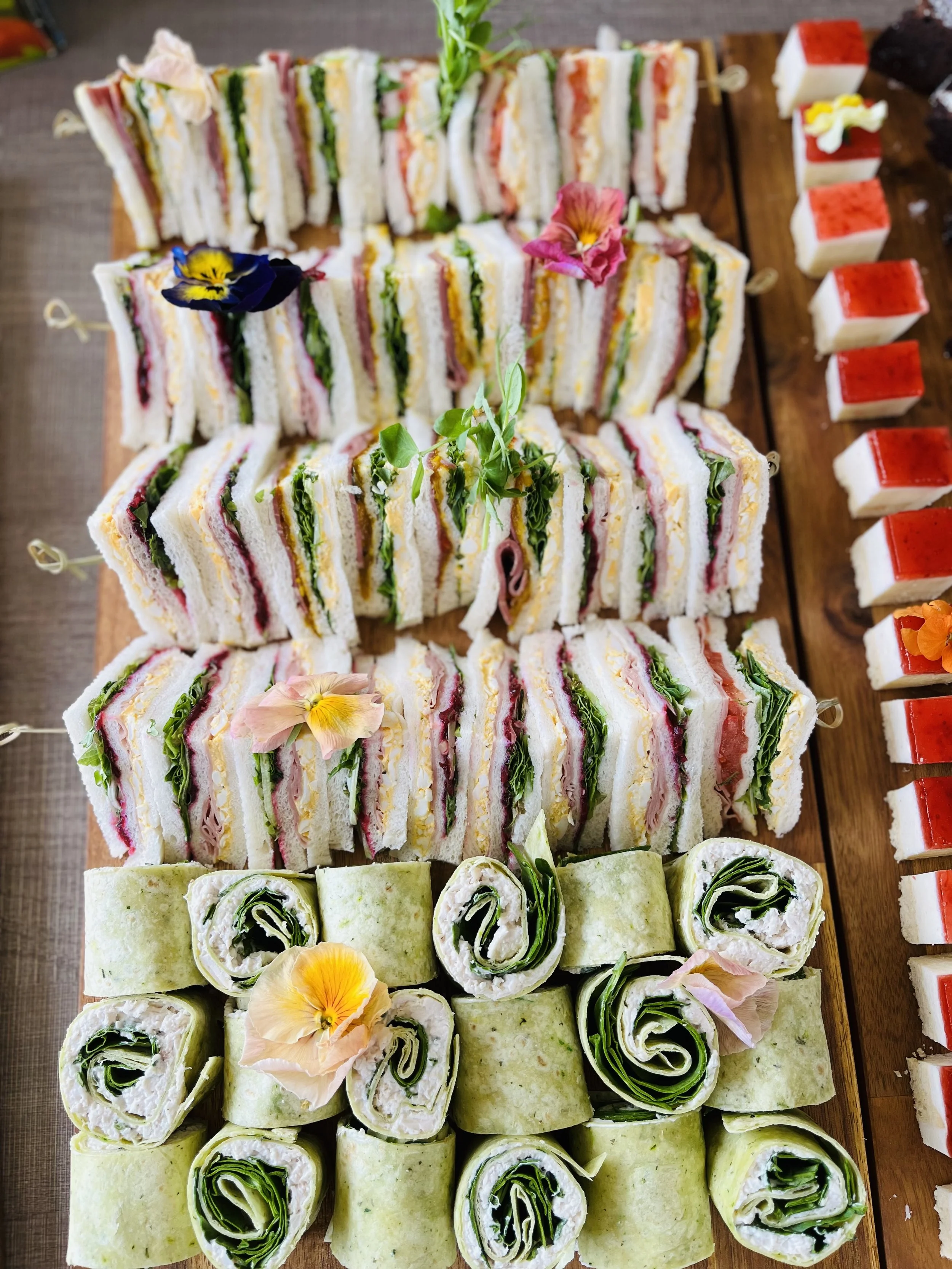 Assorted mini sandwiches and wraps arranged on a wooden platter, garnished with edible flowers.