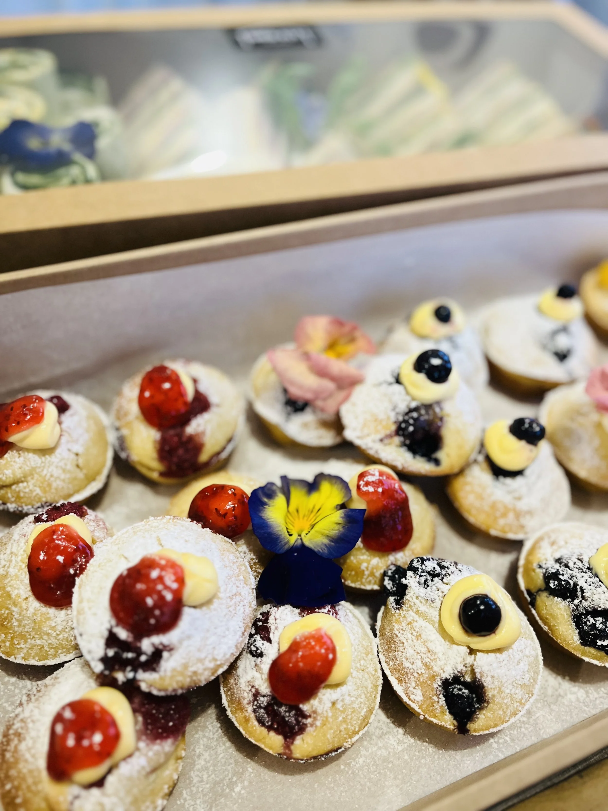Assorted mini desserts decorated with berries, edible flowers, and powdered sugar on a tray at a bakery or dessert table.