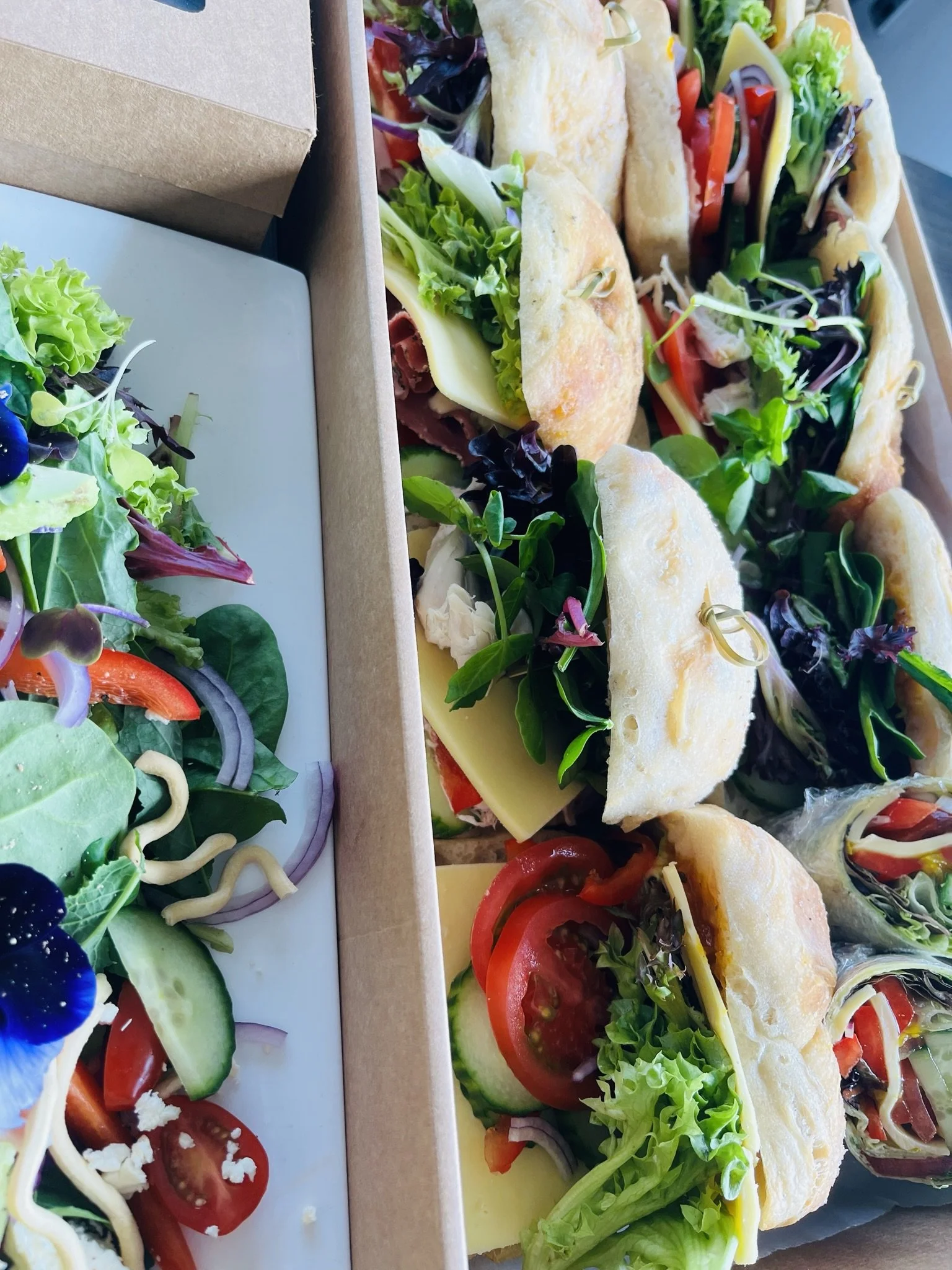 A cardboard box filled with small sandwiches stuffed with leafy greens, tomatoes, cheese, and deli meat, with some on a plate with a side salad containing mixed greens, cucumbers, cherry tomatoes, and purple onion slices.