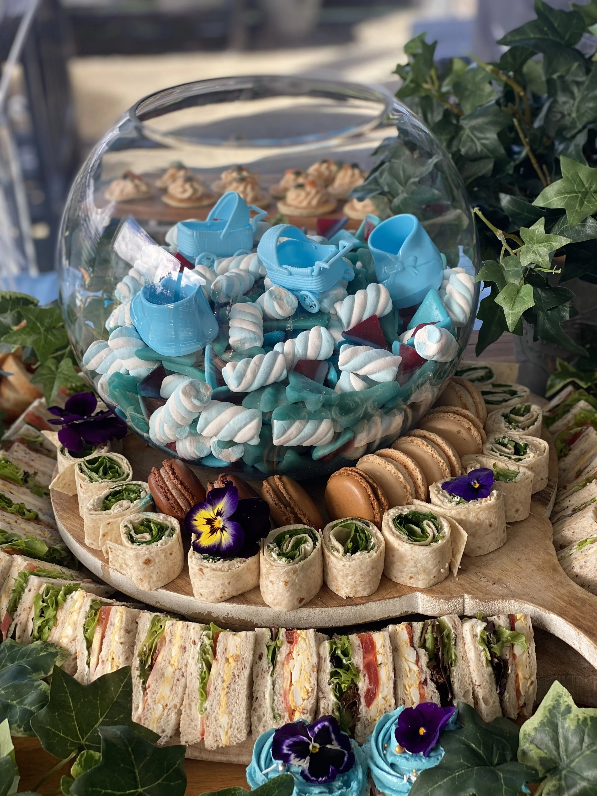 A wooden platter with assorted finger sandwiches, macarons, and rolled wraps, surrounded by flowers and greenery. In the center, a glass bowl filled with candies and small blue toy baby strollers.