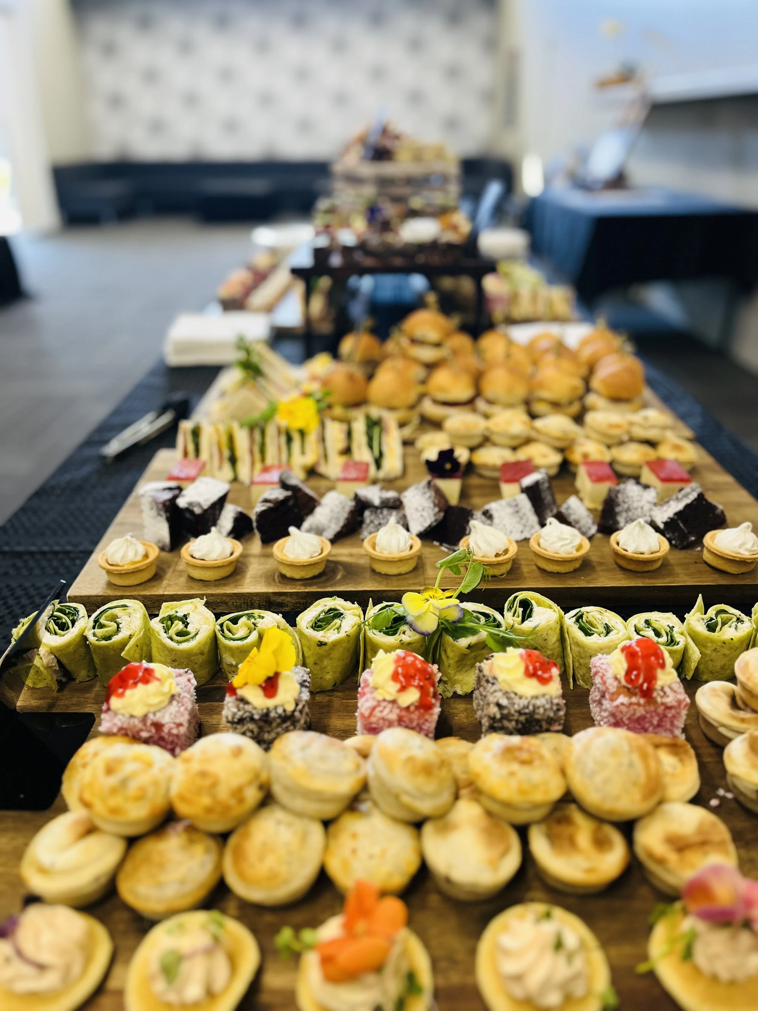 A variety of assorted finger foods and desserts arranged on a table, including mini sandwiches, tartlets, sushi rolls, and cake squares, with a blurred background.