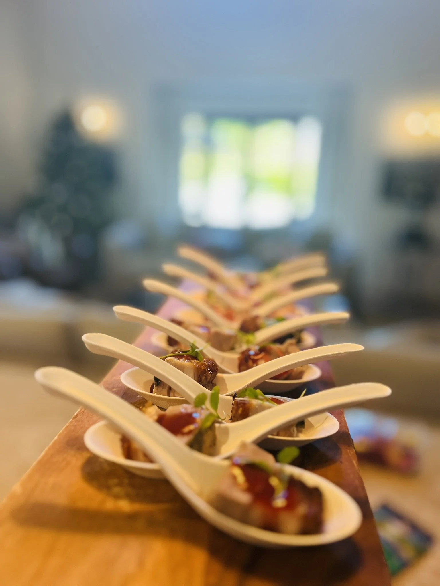 Several small appetizers served on white spoons on a wooden platter with a blurred indoor background.