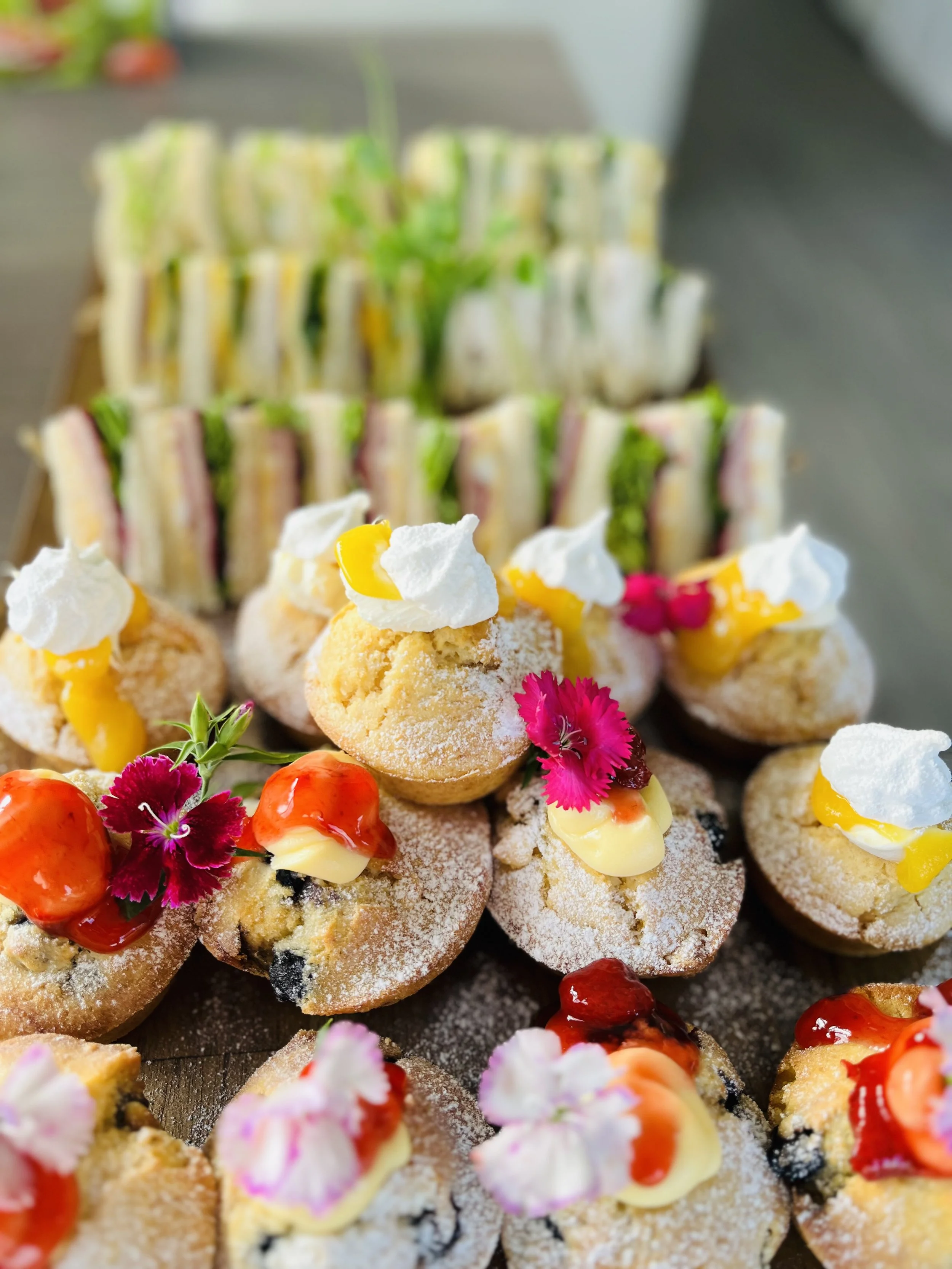 Assorted decorated pastries, including cream-filled cupcakes with pink and purple flowers, and finger sandwiches including cucumber, tomato, and ham on white bread with various fillings.