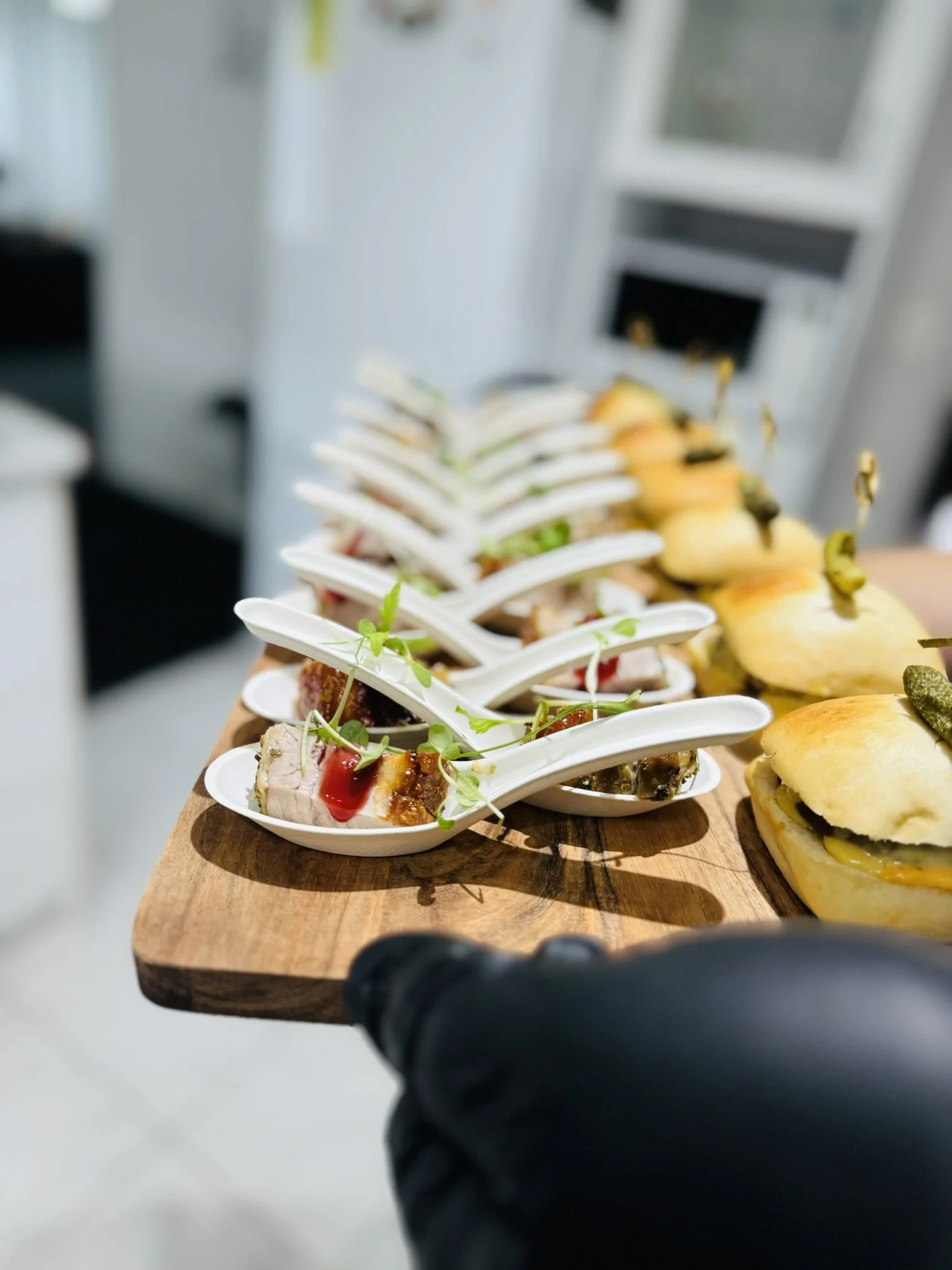 A wooden serving board with small white porcelain spoons containing gourmet appetizers, including meats with sauces and garnishes, and mini sandwiches or sliders with pickles.