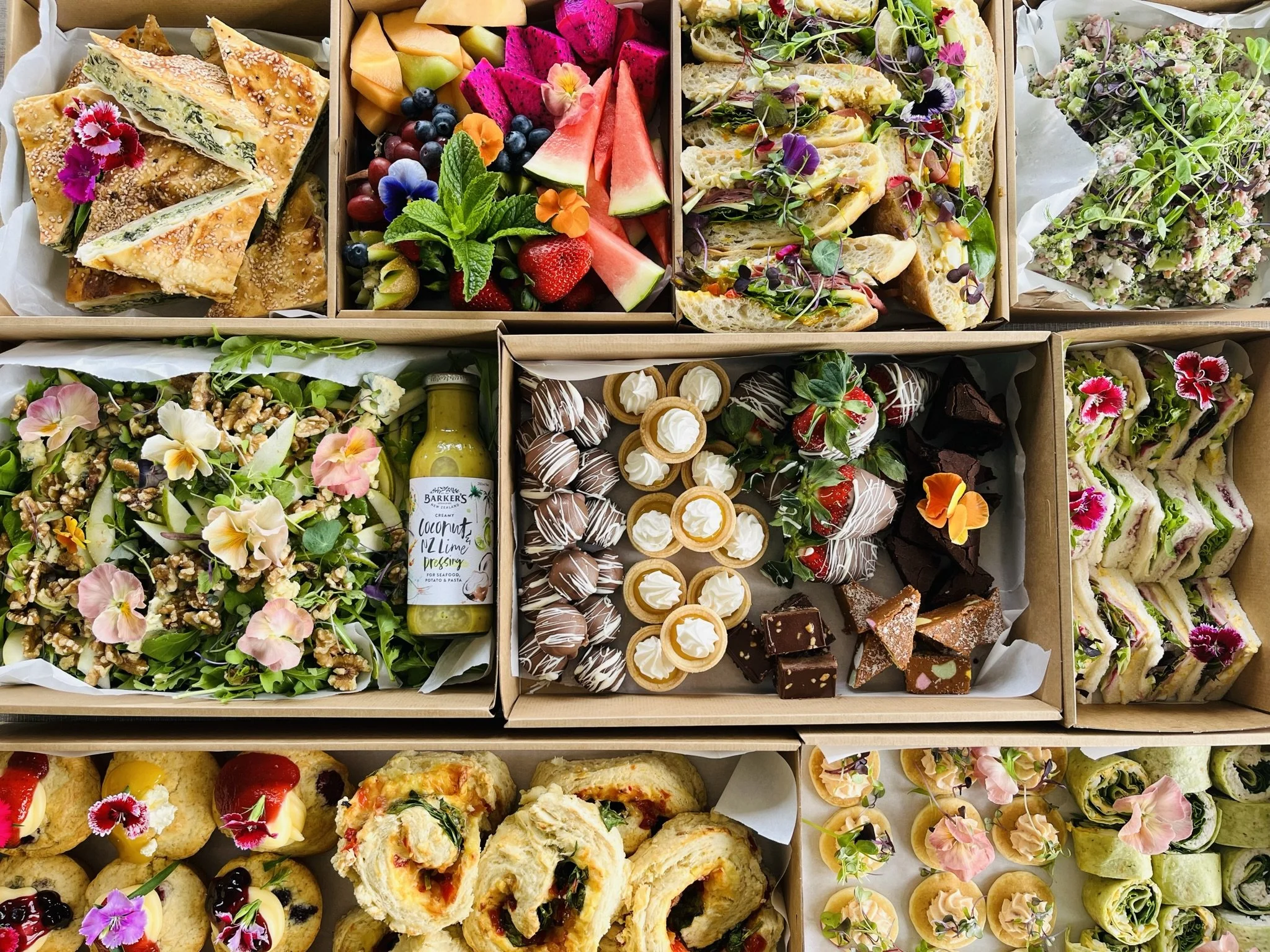 An assortment of colorful sandwiches, salads, fresh fruit, and desserts arranged in boxes, including fruit skewers, chocolate truffles, mini tarts, and savory pinwheels, decorated with edible flowers.