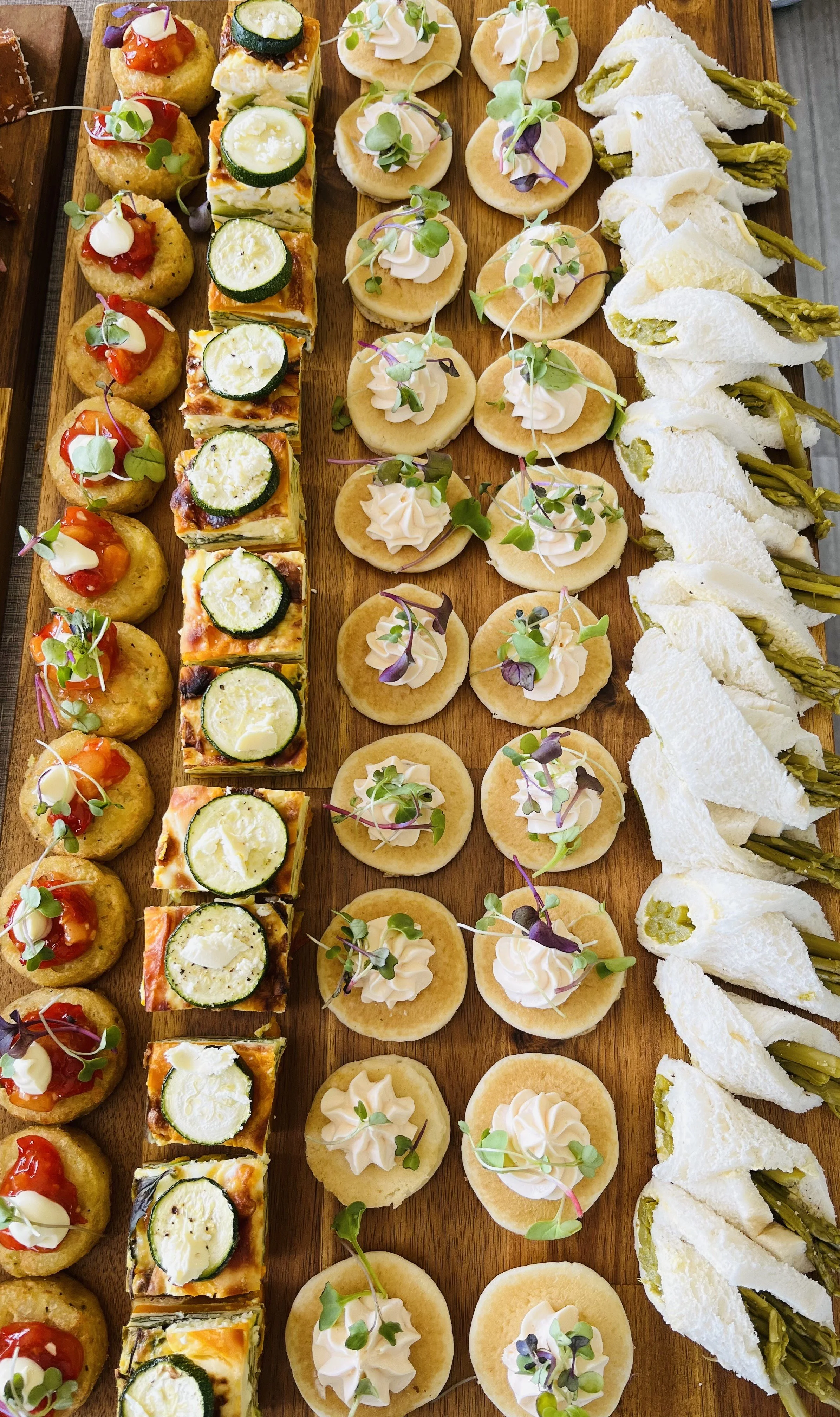 Assorted canapé appetizers arranged on a wooden platter, including mini potato bites with cherry tomatoes and microgreens, zucchini and cheese tartlets, round crackers topped with cream and microgreens, and white bread sandwiches with green filling.
