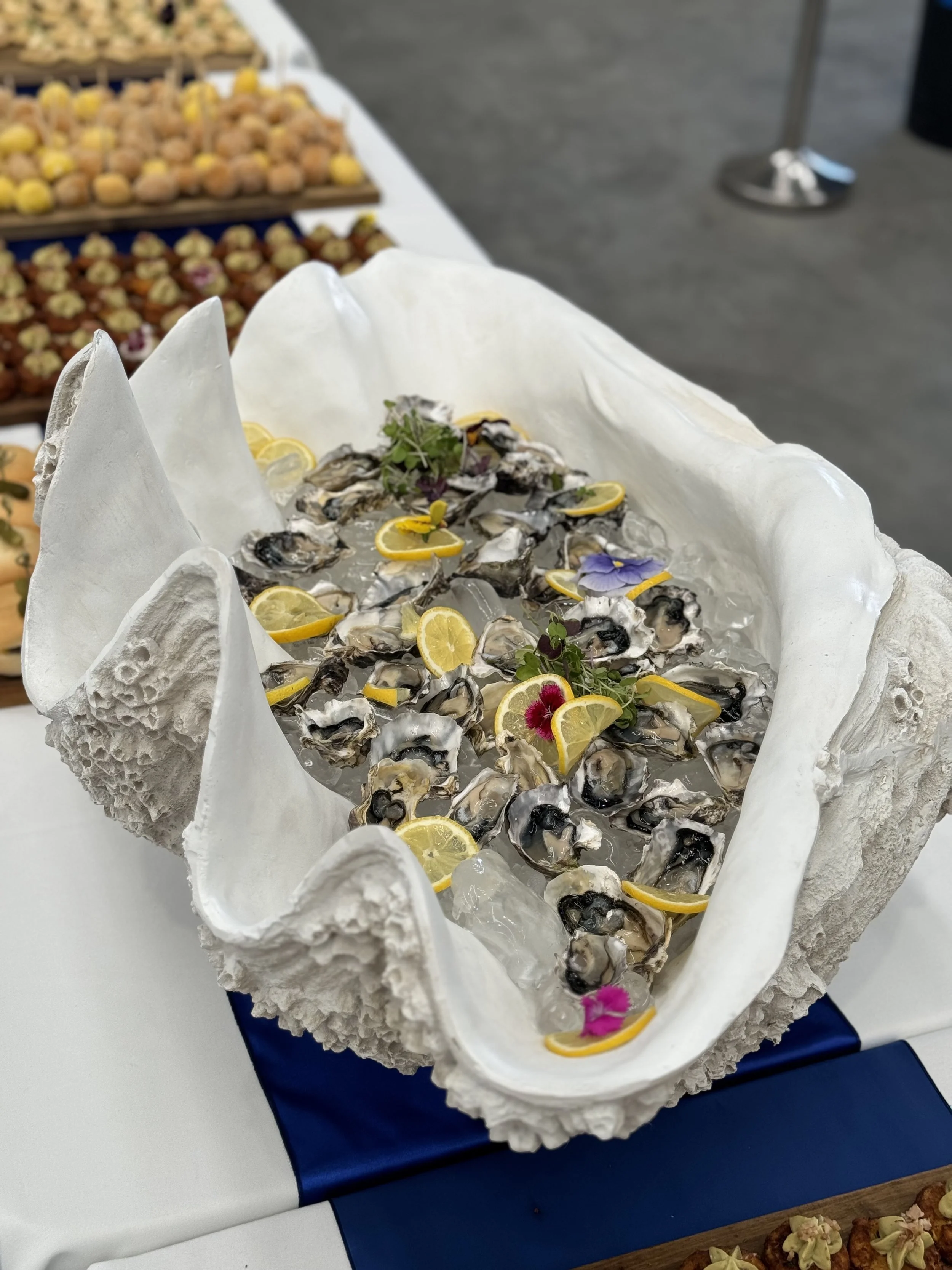 A seafood platter with oysters topped with lemon slices and edible flowers on ice, in a large white shell-shaped container.
