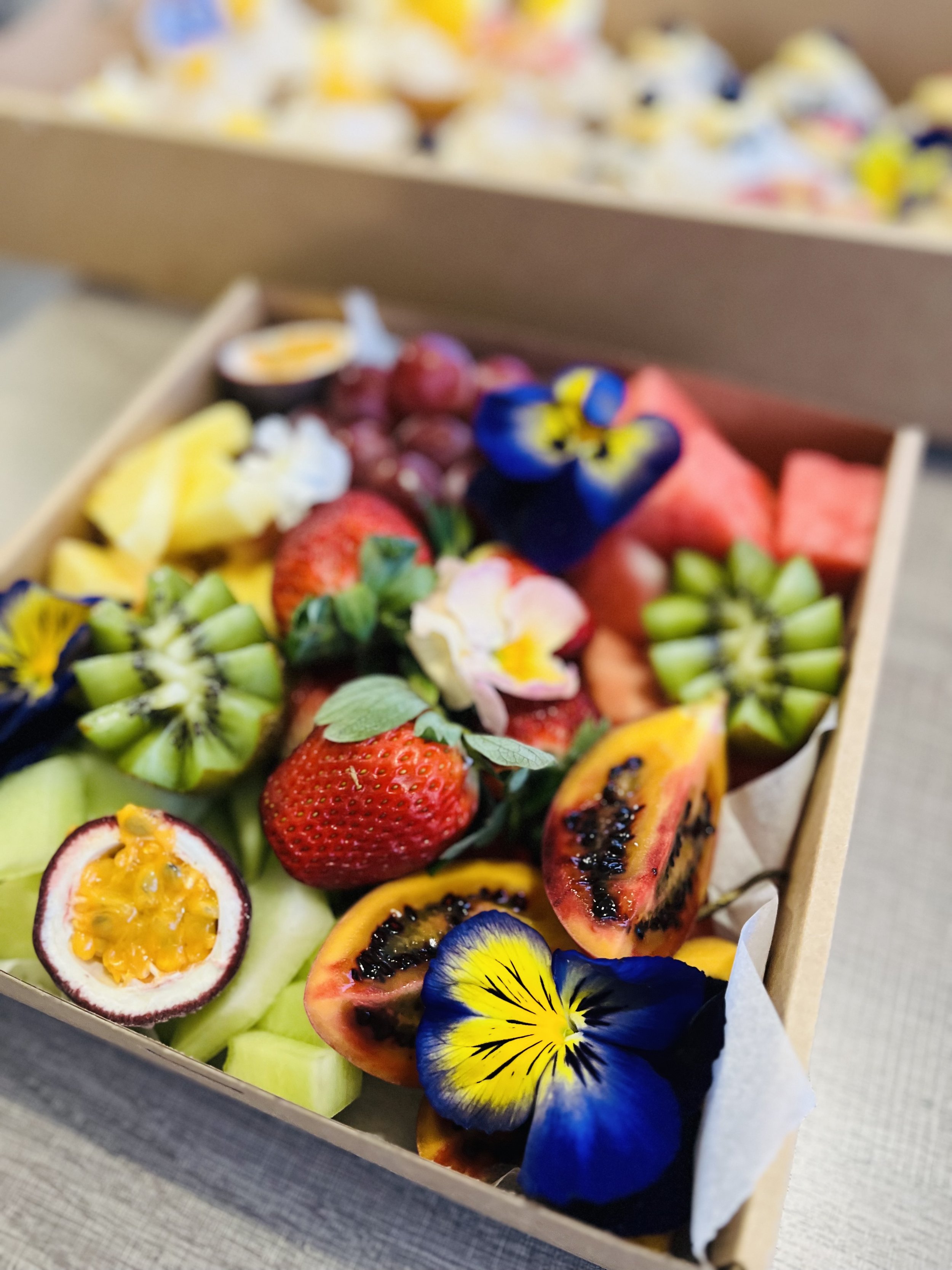 A wooden box filled with assorted fresh fruits including strawberries, passion fruit, kiwi, watermelon, and edible flowers.