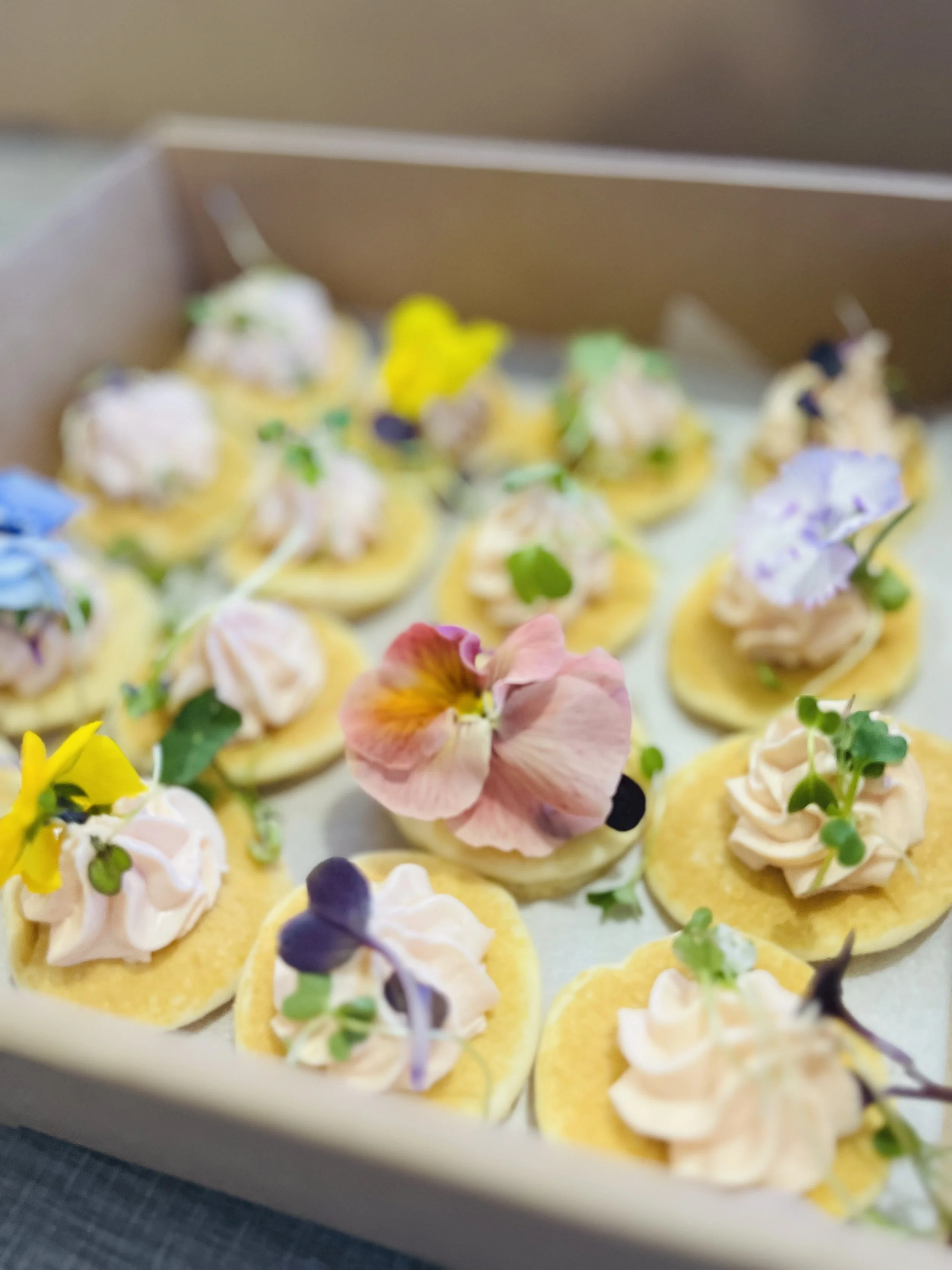 Assorted gourmet cupcakes decorated with sprigs of herbs and edible flowers, placed in a cardboard box.