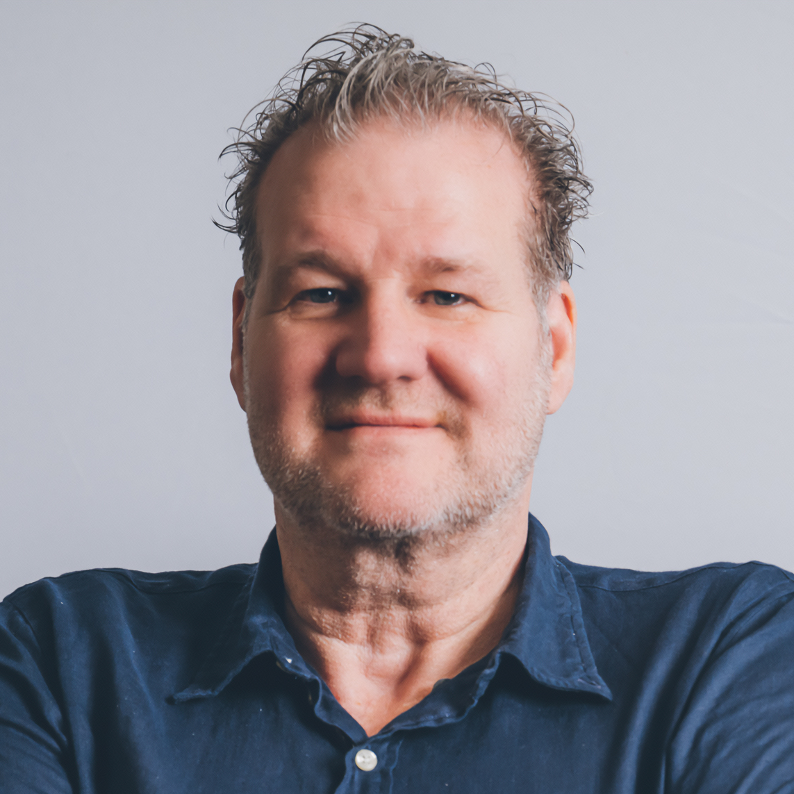 A smiling middle-aged man with short, slightly curly hair and a beard, wearing a dark blue button-up shirt, standing against a plain light gray background.