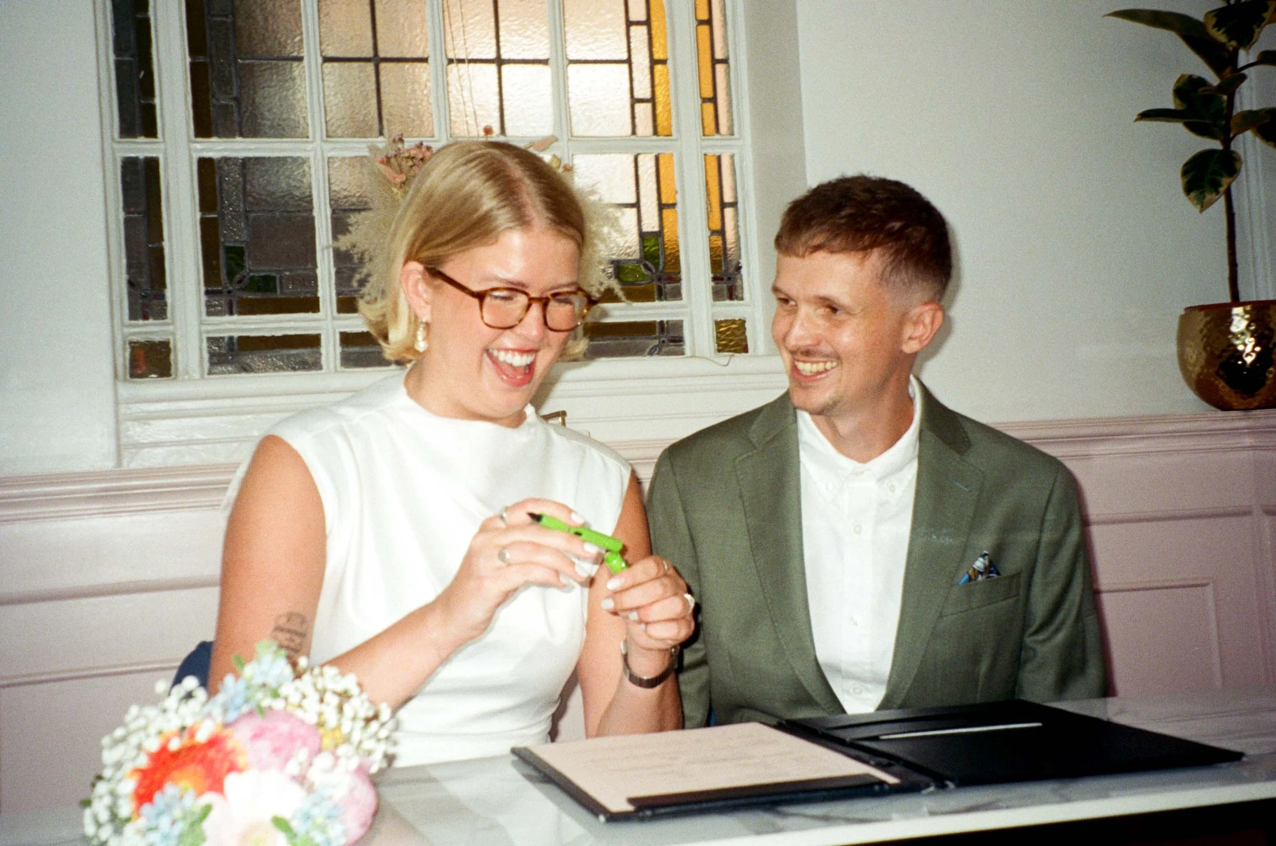 A woman with blonde hair and glasses, dressed in a white sleeveless dress, is smiling and looking at her hands, possibly opening a gift. A man in a green suit is sitting beside her, also smiling and looking at her. There are flowers on the table in front of them and a decorative window with stained glass in the background.