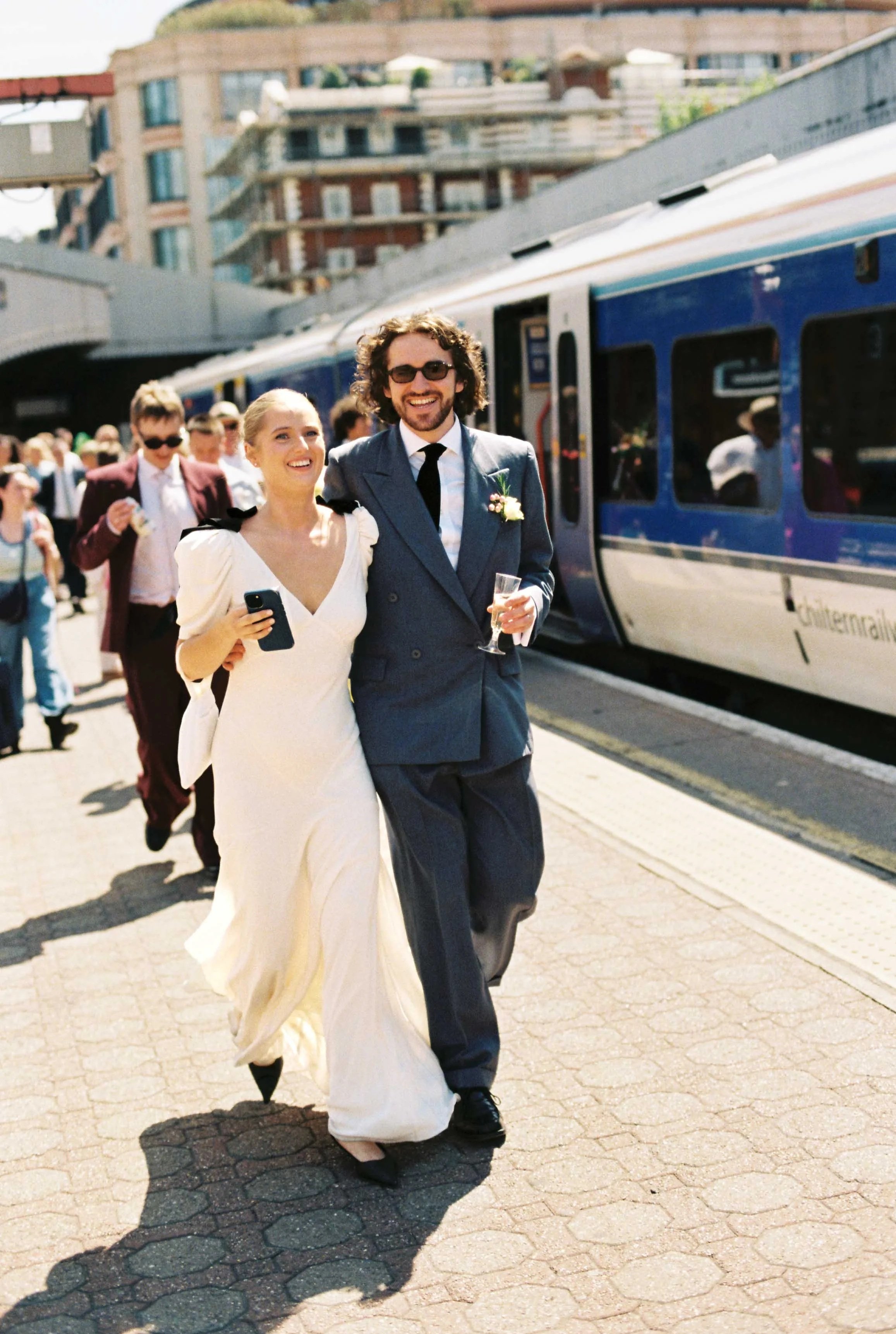 A newlywed couple walking together at a train station, smiling and laughing. The bride is wearing a white wedding gown and holding a phone, and the groom is in a blue suit with sunglasses and holding a glass of champagne. There are other people in the background, and a blue train is visible beside them.