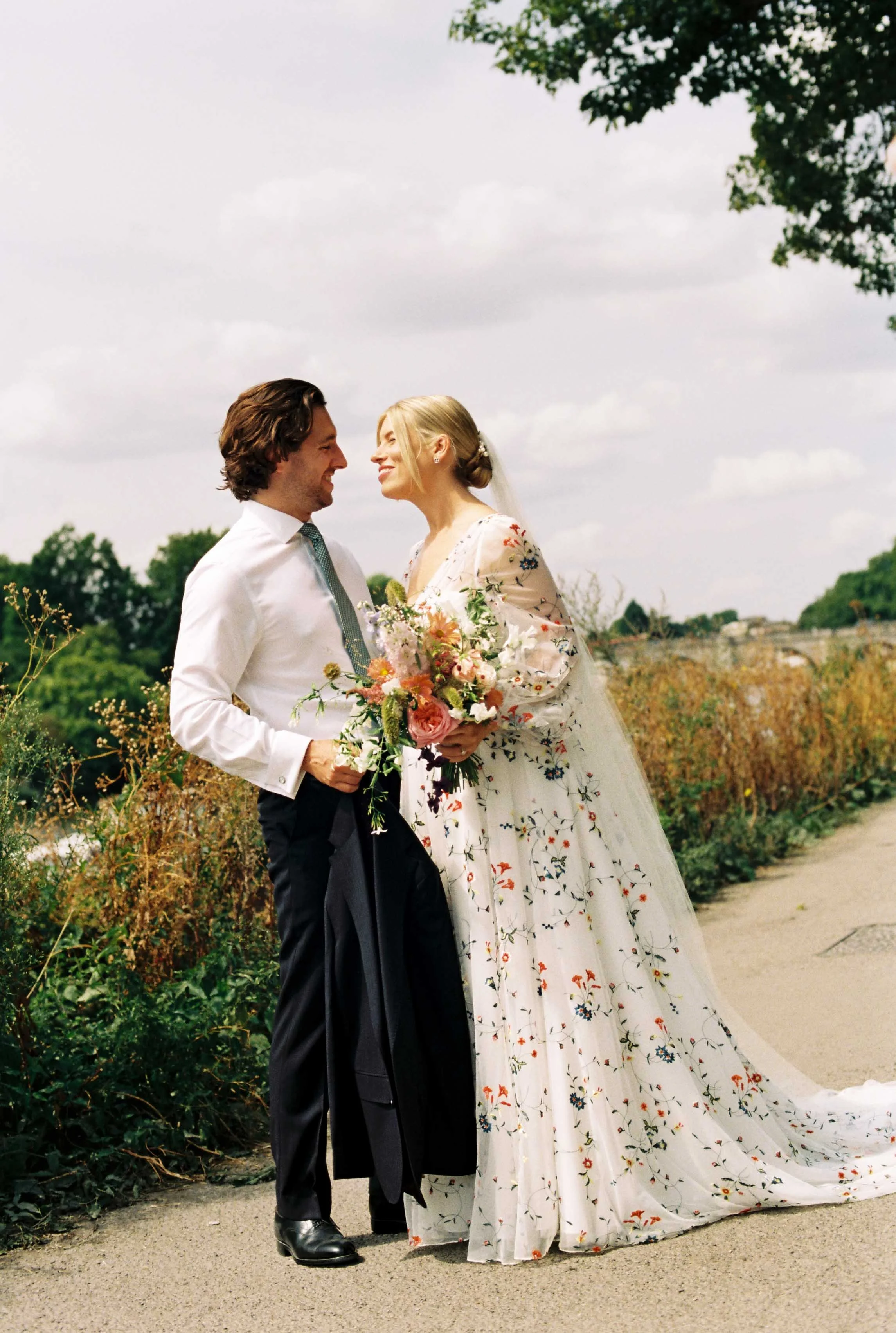 A couple dressed in wedding attire standing outdoors, smiling and looking at each other, with a natural landscape and cloudy sky in the background.