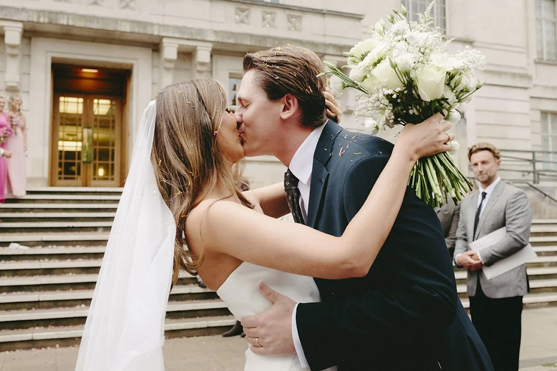 A bride and groom kiss in front of a building on their wedding day, with the bride holding a bouquet of white flowers and the groom wearing a navy blue suit.