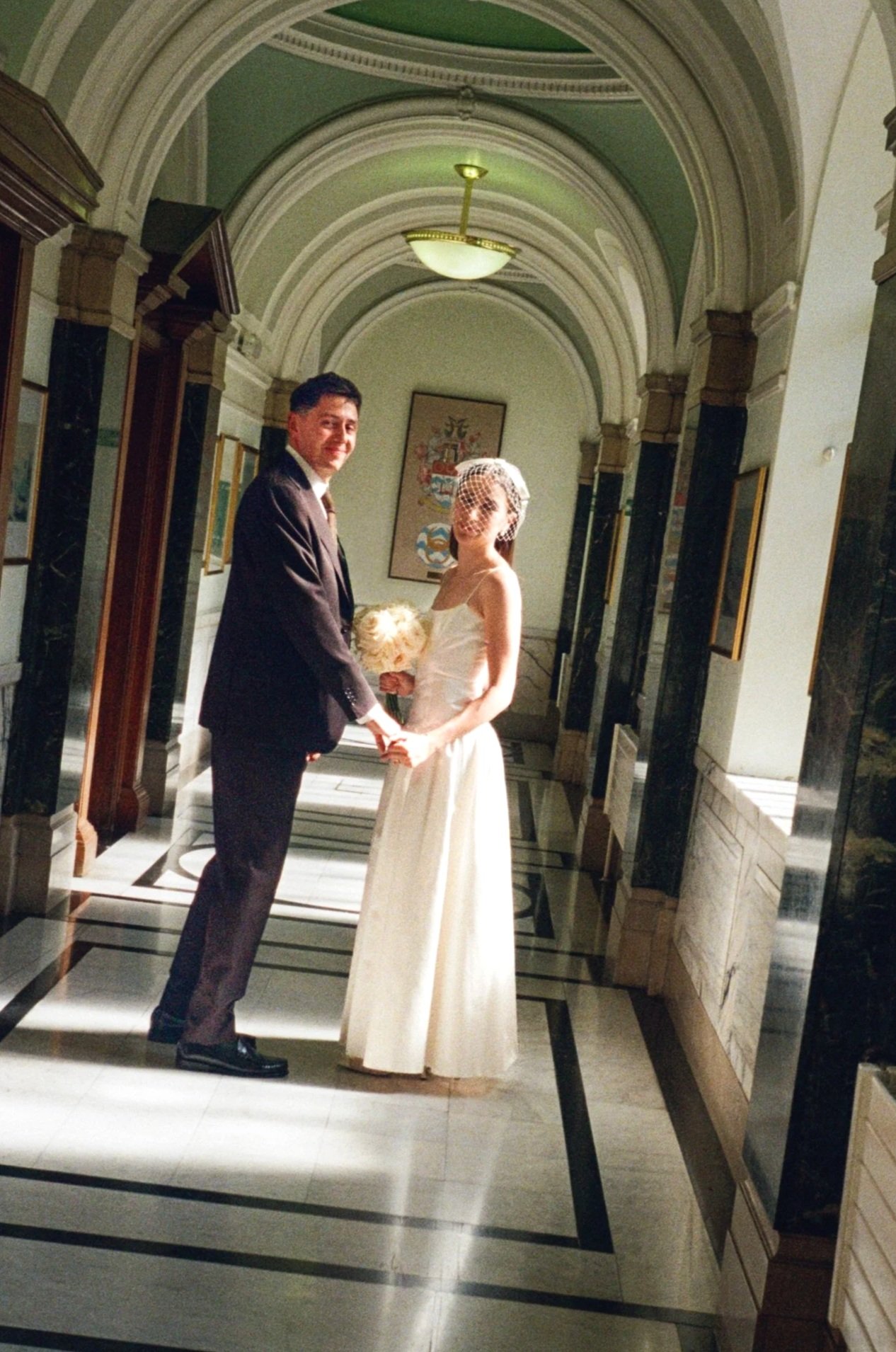 A man and woman holding hands in formal attire in a hallway with arched ceilings and framed artwork on the walls.
