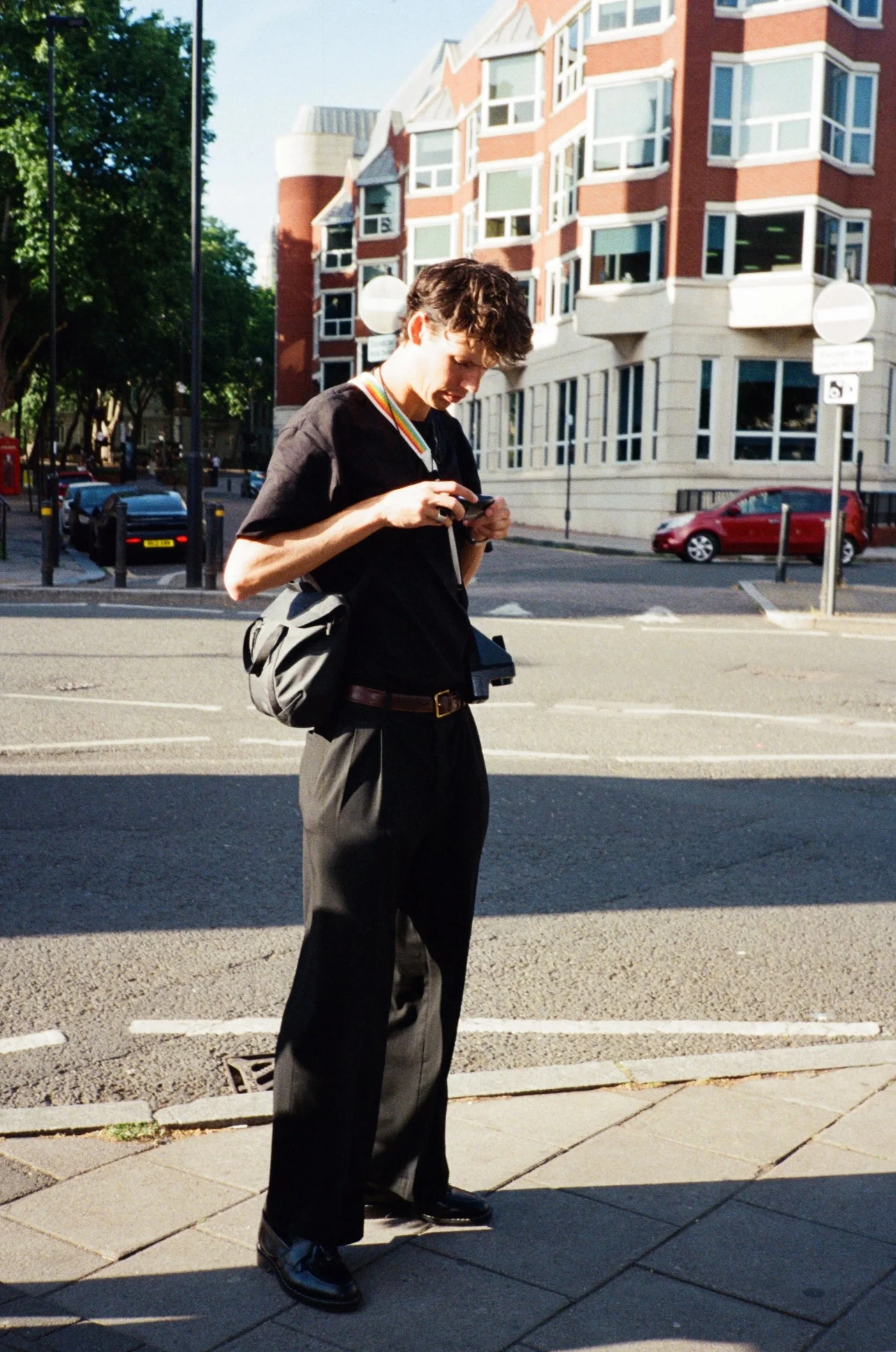 A man with dark, curly hair looking down at his phone while standing on a city sidewalk. He is wearing a black shirt and dark trousers, carrying a backpack. The background features a multi-story brick building with large windows and parked cars along the street, under a clear sky.