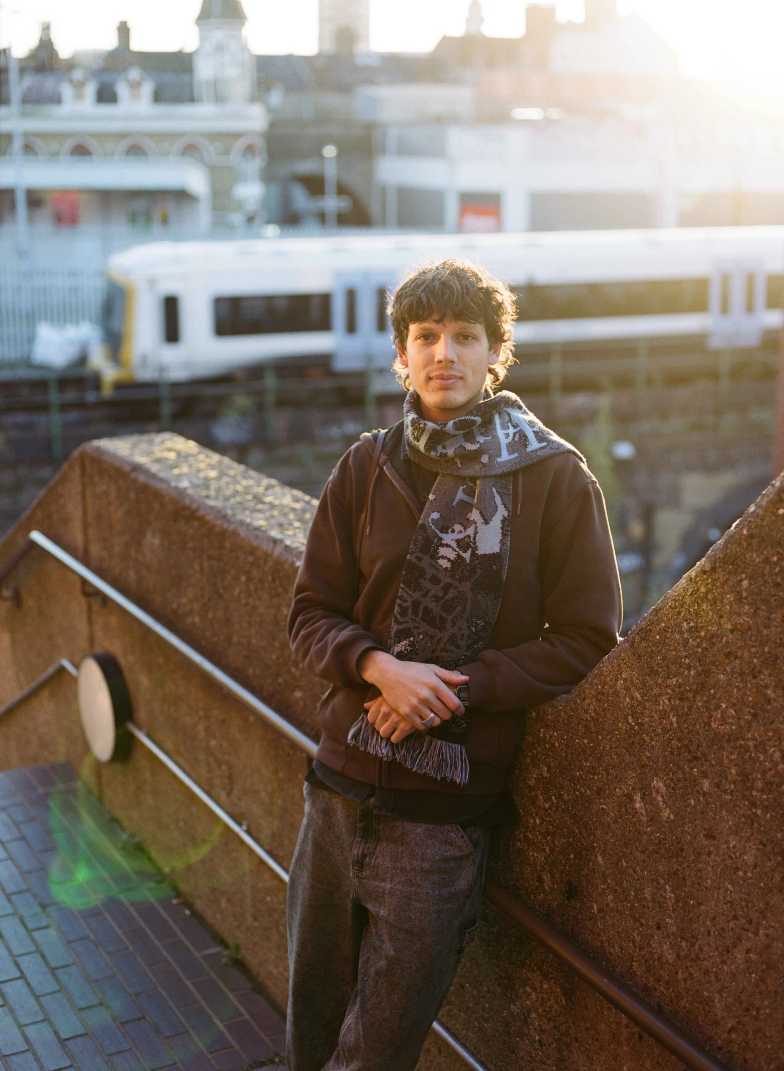 A young man with dark wavy hair and a scarf around his neck standing outdoors on a bridge, with a cityscape and a white train in the background during sunset.