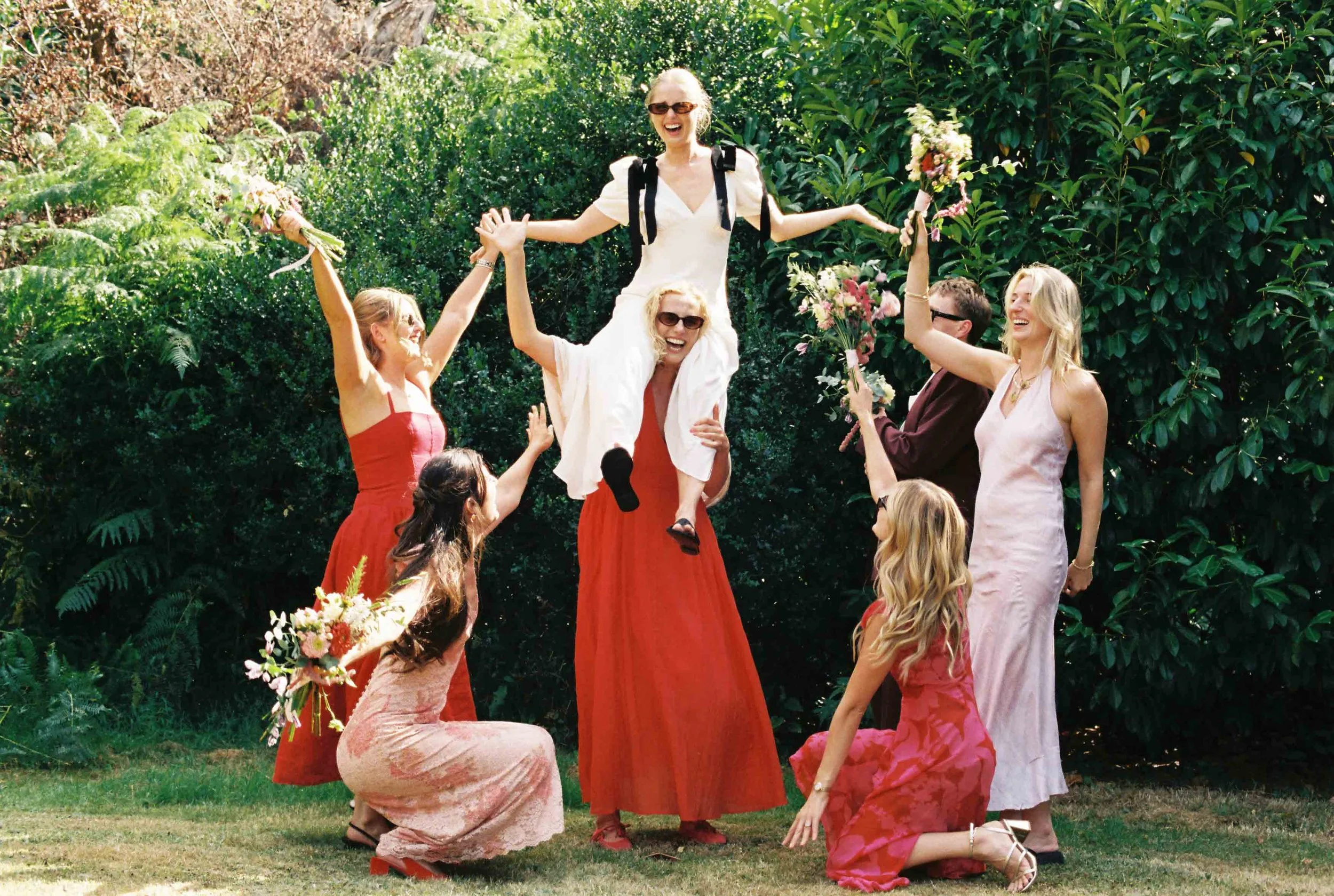Group of women celebrating outdoors, with one woman being lifted by others, holding flowers, all smiling, surrounded by greenery.