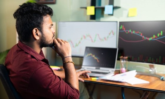 A young man in an office, looking at computer screens showing stocks-related stats.