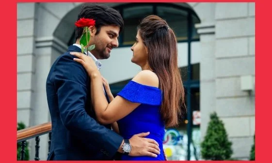 A man with is his hands on a woman’s waist, and the woman’s hands on his shoulders holding a flower.