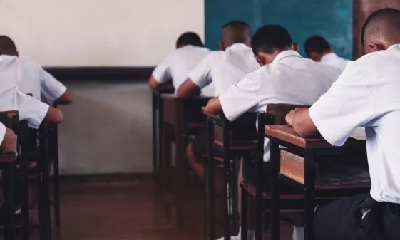 A classroom, with students shown from the back writing something at their desks.