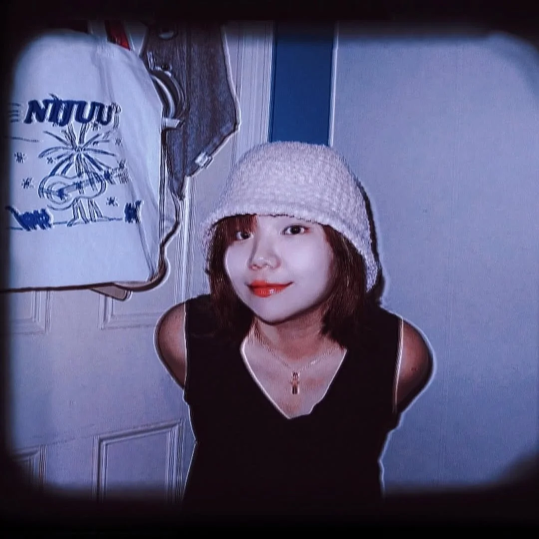 Person wearing a white knit hat and black top, standing indoors near a hanging bag with an illustration and text.