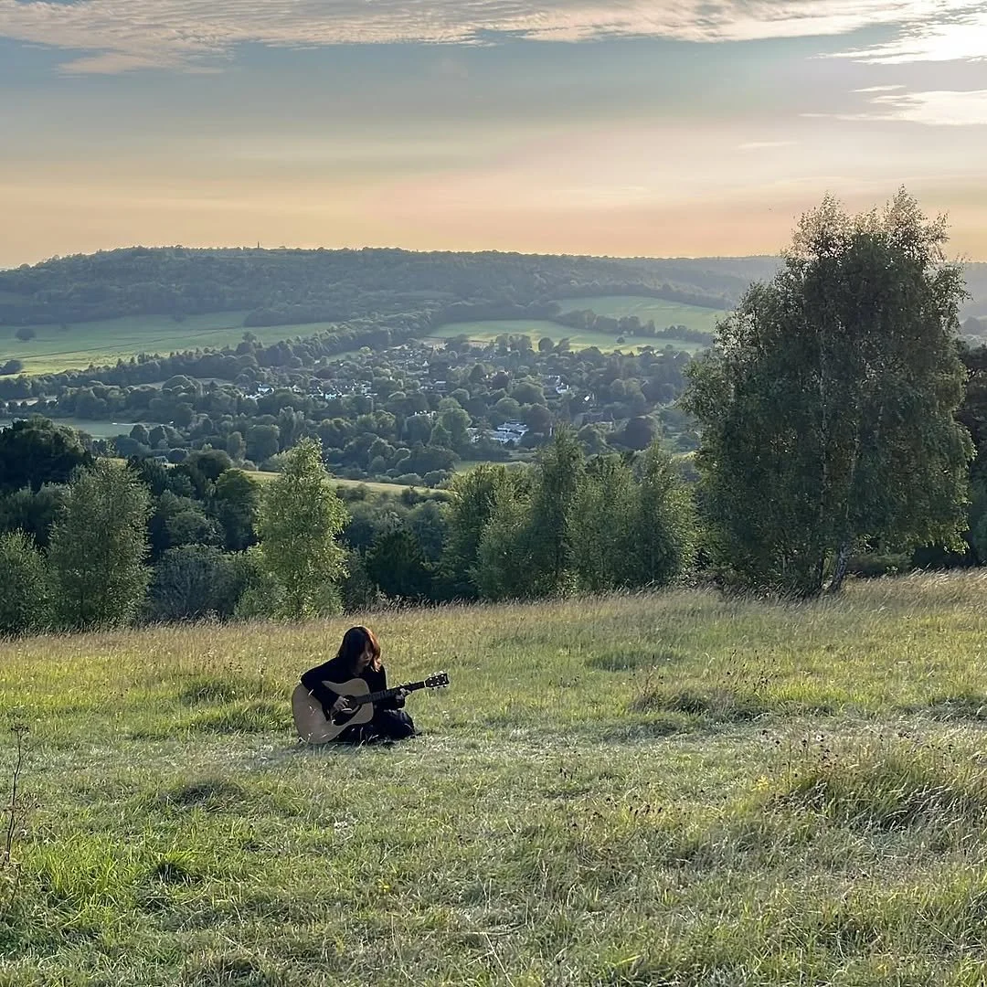 Person sitting in a grassy field playing a guitar with hills and a small town in the background under a partly cloudy sky.
