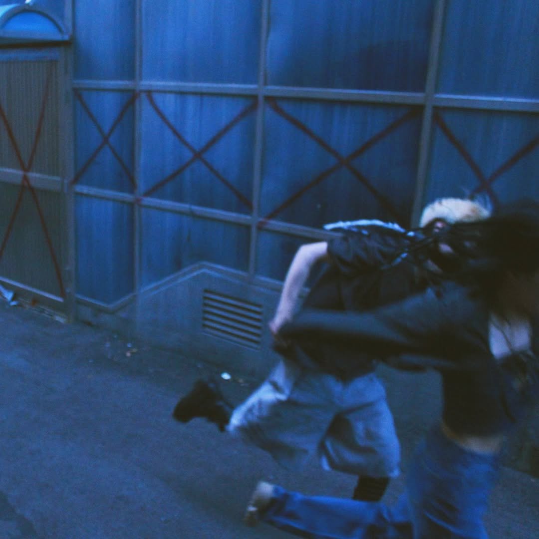 Two people running in a dimly lit alley with blue walls and graffiti markings.