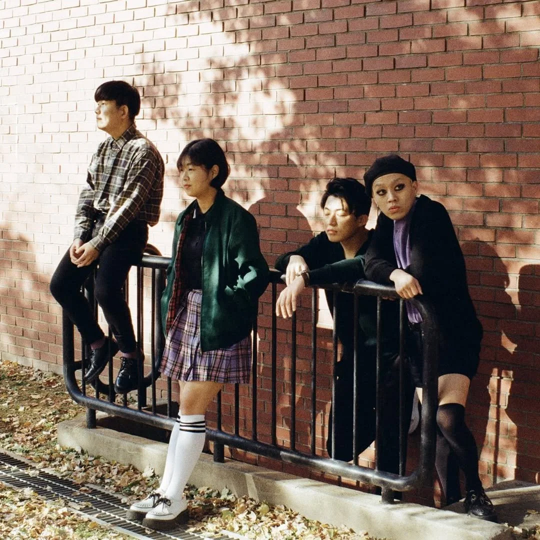 Four people standing and sitting by a brick wall, wearing casual fall clothing and leaning on a black metal rail.