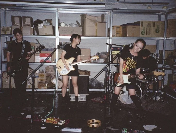 Four musicians performing in a storage room, with two guitarists and a bassist in the front and a drummer in the back. Musical equipment and boxes visible in the background.