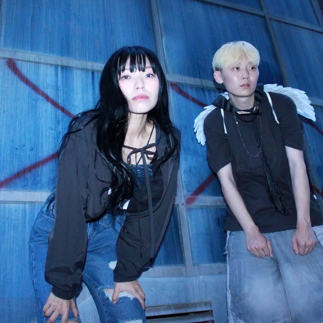 Two individuals in casual clothing stand in front of a blue textured wall with red graffiti. One has long black hair and the other has short blond hair with angel wings. The mood is edgy and urban.