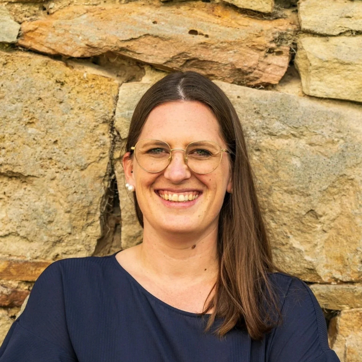 Eine lachende Frau mit Brille vor einer Steinfeldmauer.