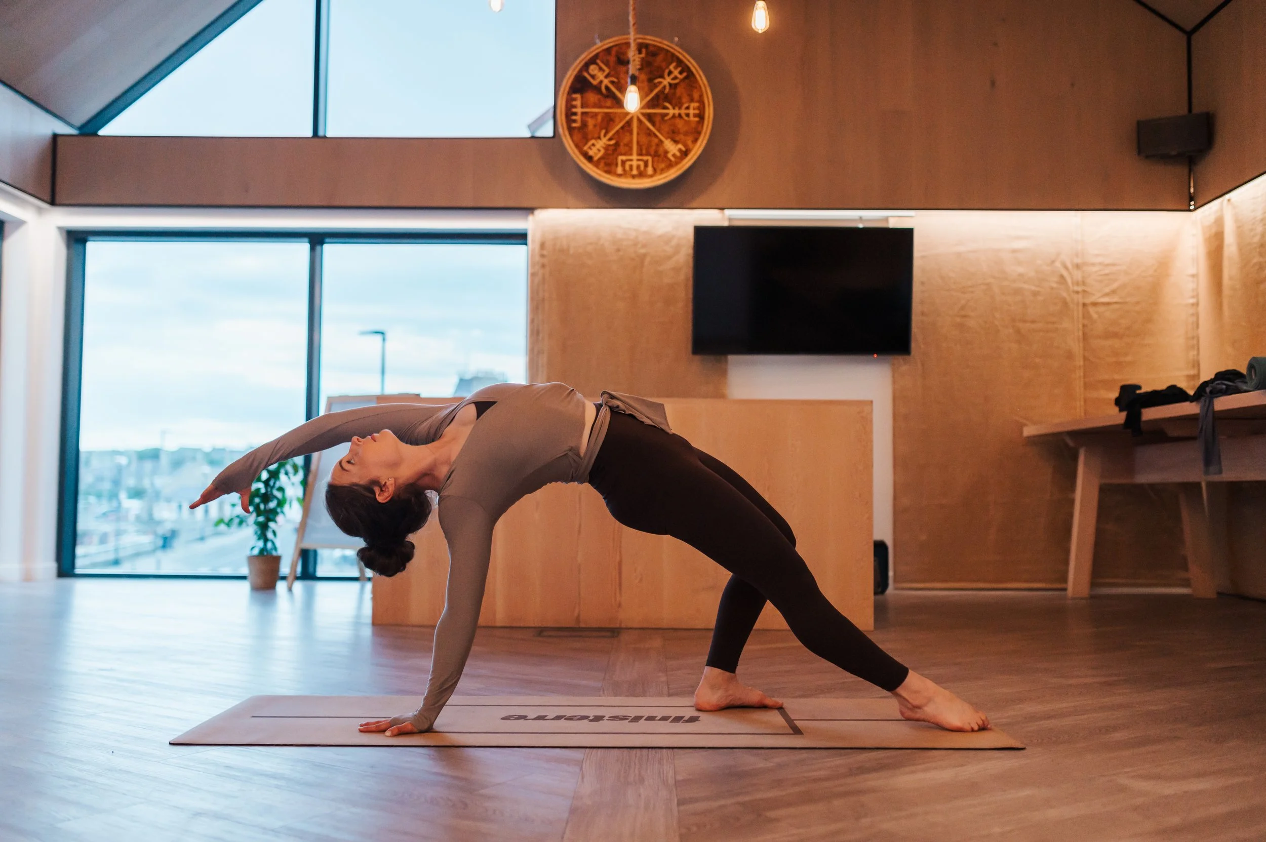 A woman practicing yoga in a spacious, modern indoor studio with large windows, wooden walls, a wall clock, and a flat-screen TV. She is in a side plank pose on a yoga mat.