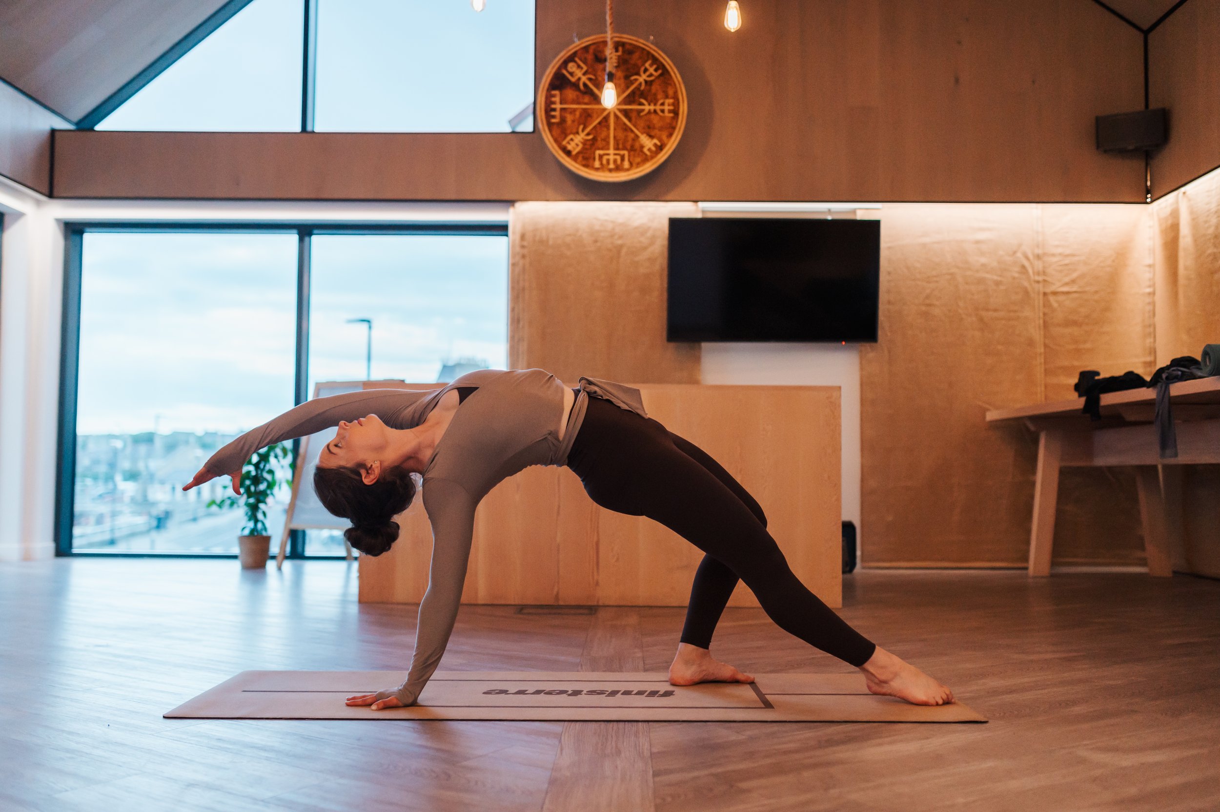 A woman practicing yoga indoors on a yoga mat, performing a side bend pose in a well-lit room with large windows, wooden walls, and a decorative clock on the wall.