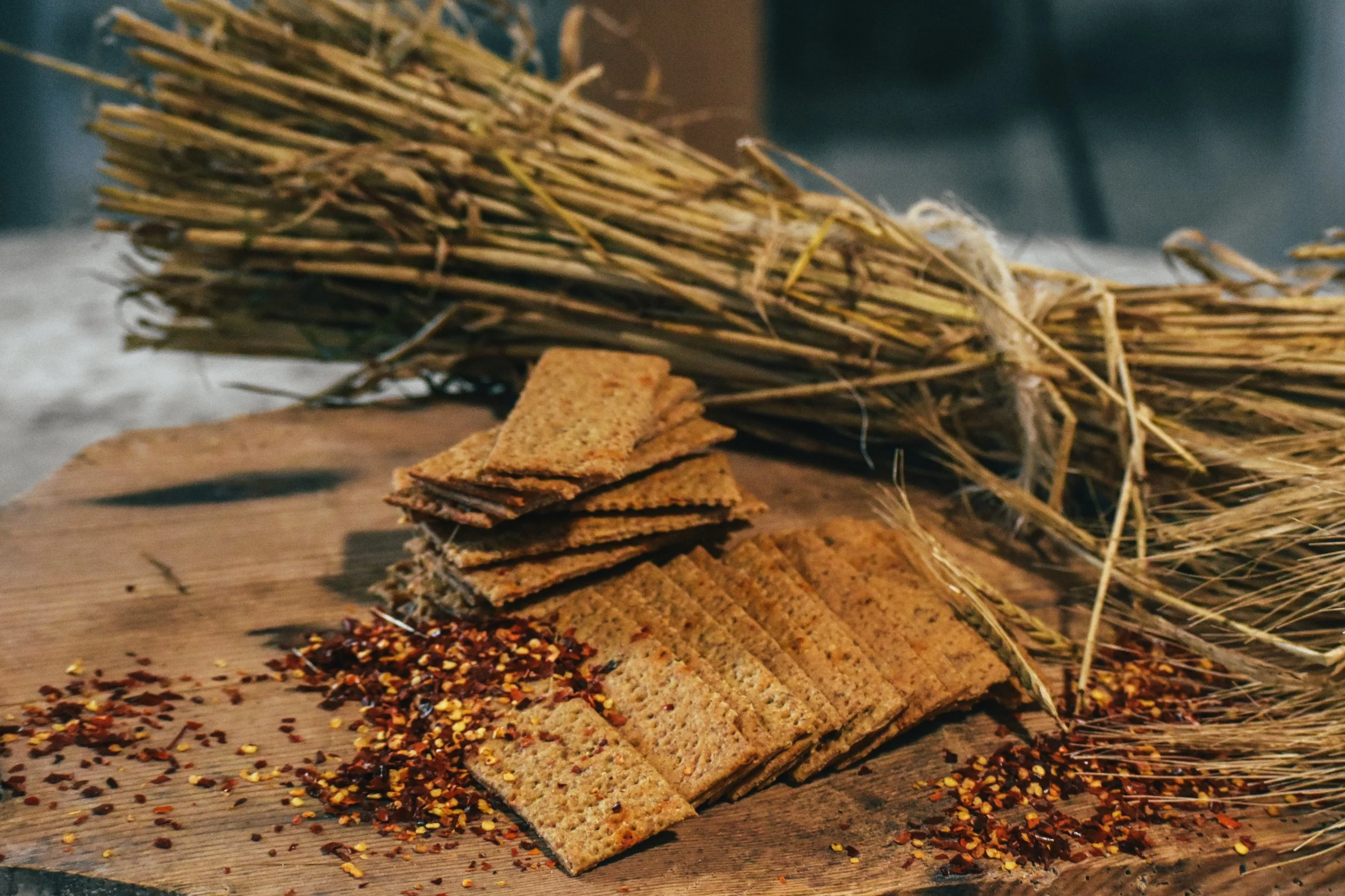 Stack of crispy crackers on a wooden surface, sprinkled with chili flakes, with a bundle of dried wheat stalks in the background.