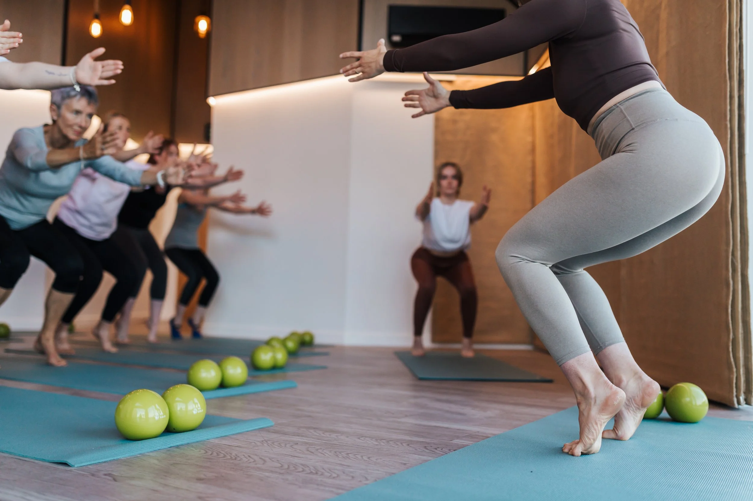 A group of women participating in a yoga class, following instructor, on yoga mats with green balls placed in rows, in a wooden-floored room with warm lighting.
