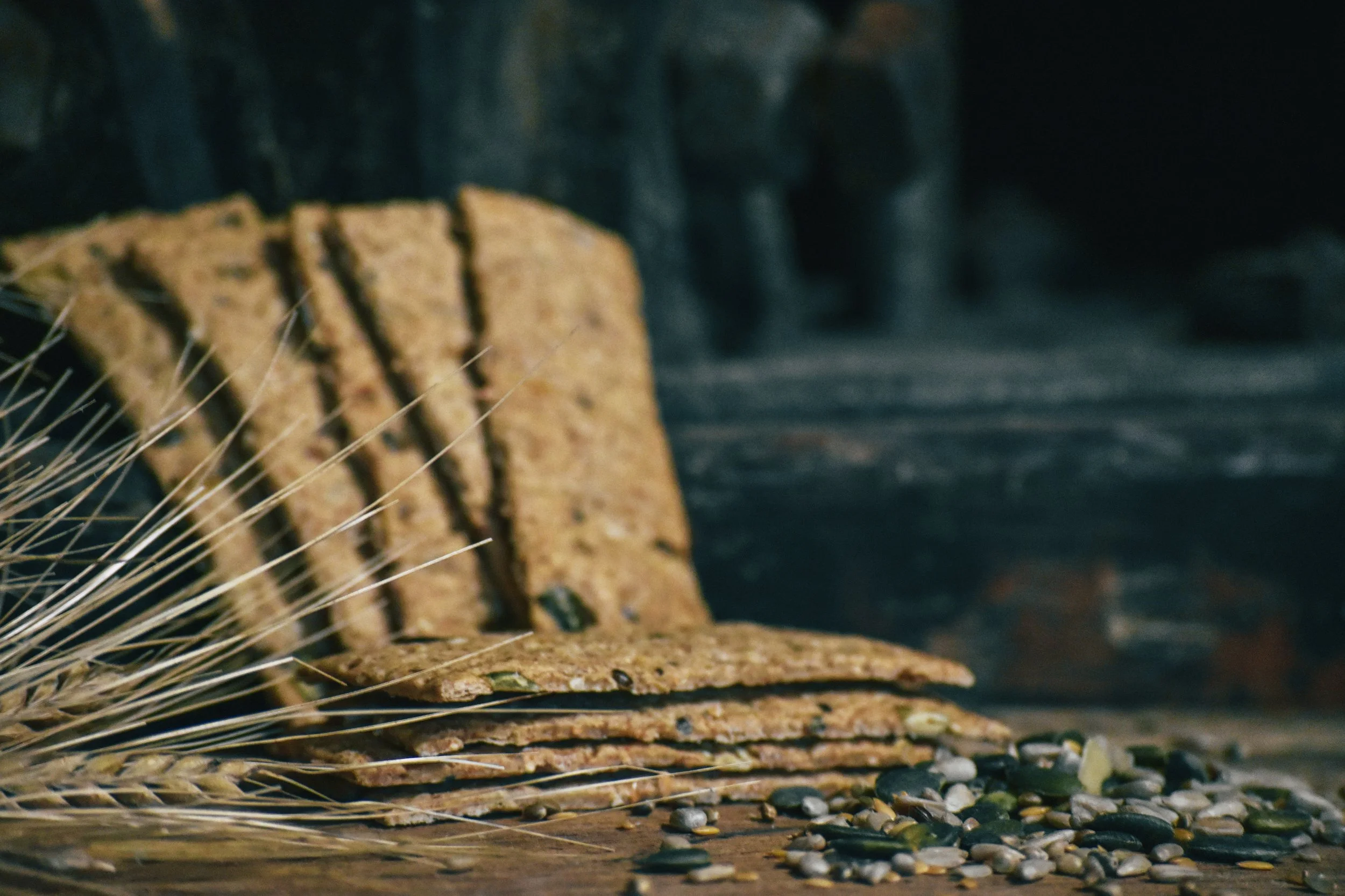 Close-up of stacked crackers and scattered seeds next to a sprig of wheat on a dark surface.