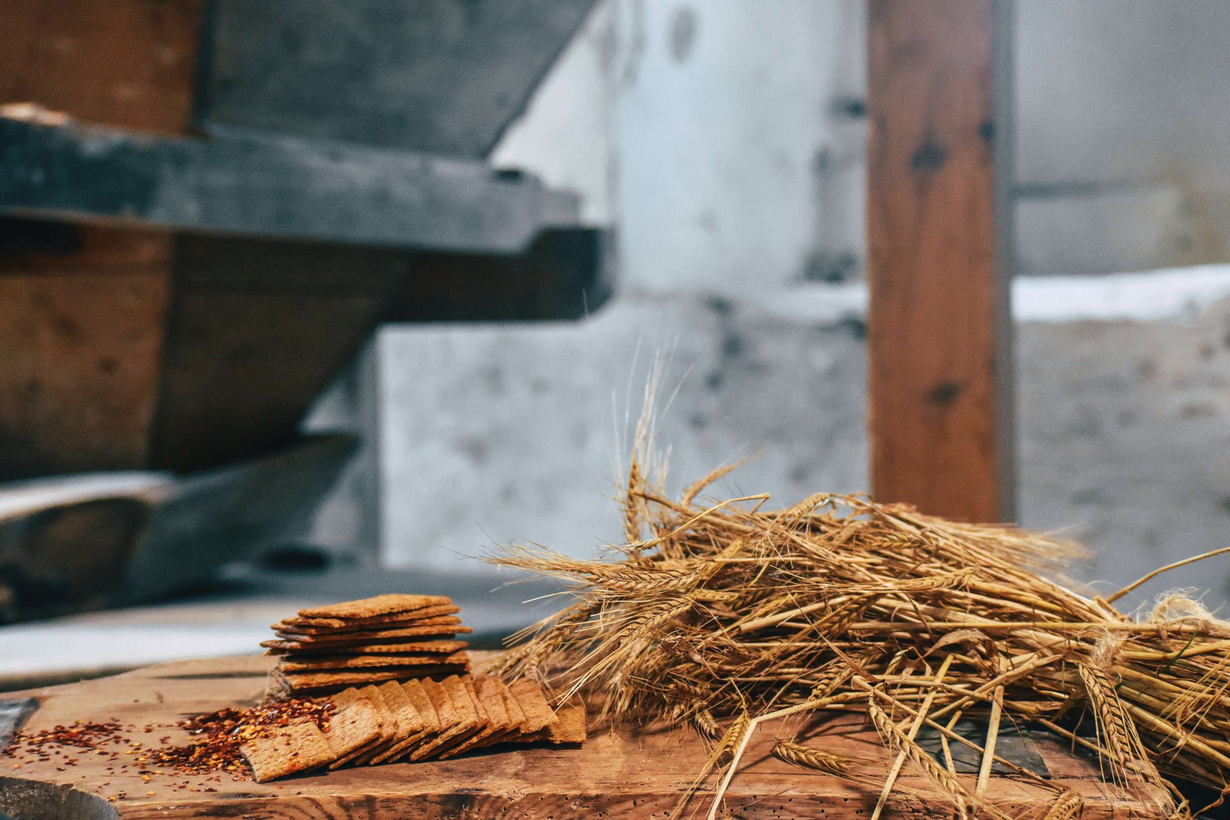 A stack of crackers, a pile of wheat stalks, and some spices on a wooden surface, with a blurry industrial background.