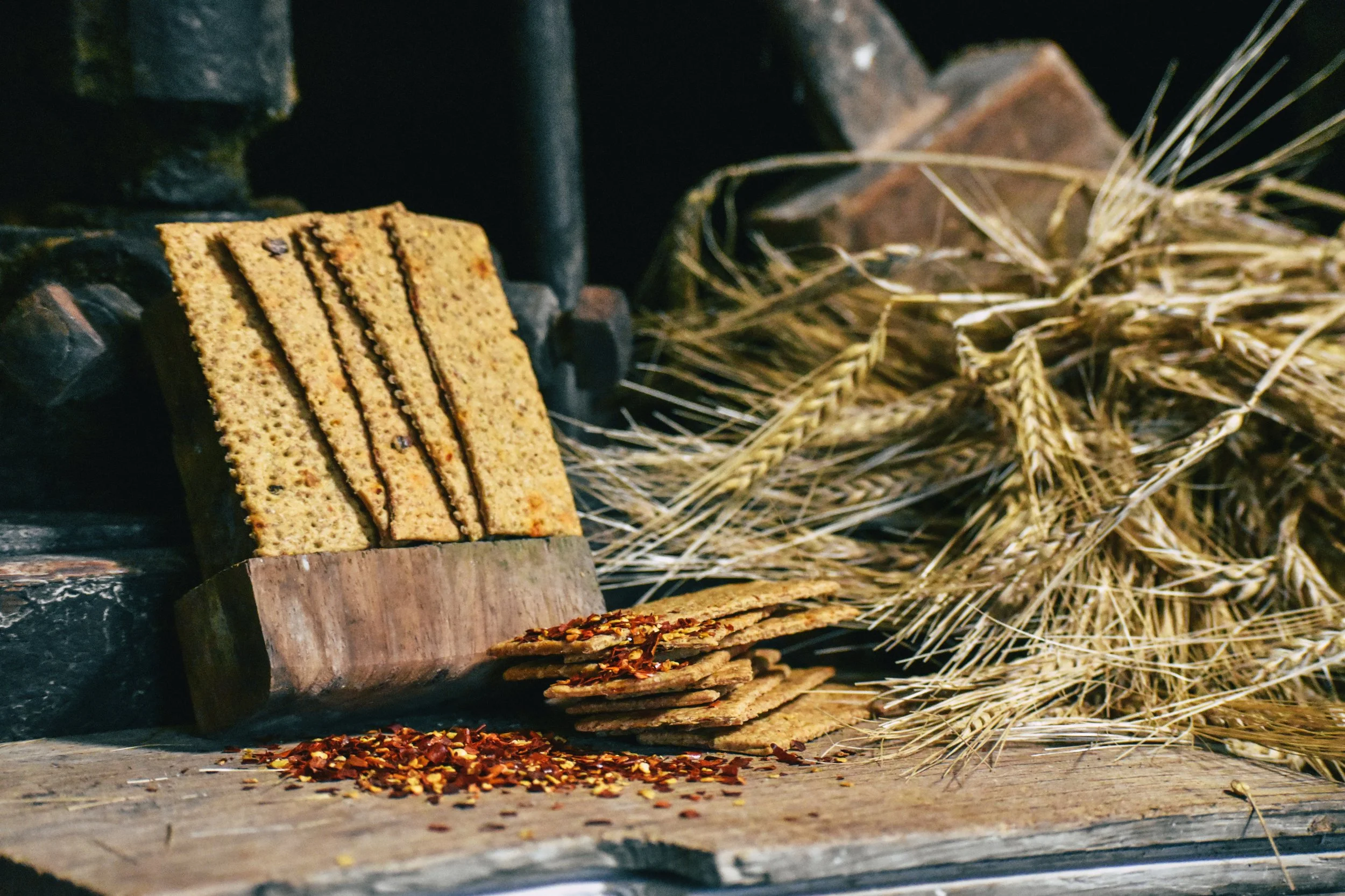 Close-up of a stack of crispbread on a wooden surface, with dried wheat and chili flakes scattered nearby, set against a rustic background.