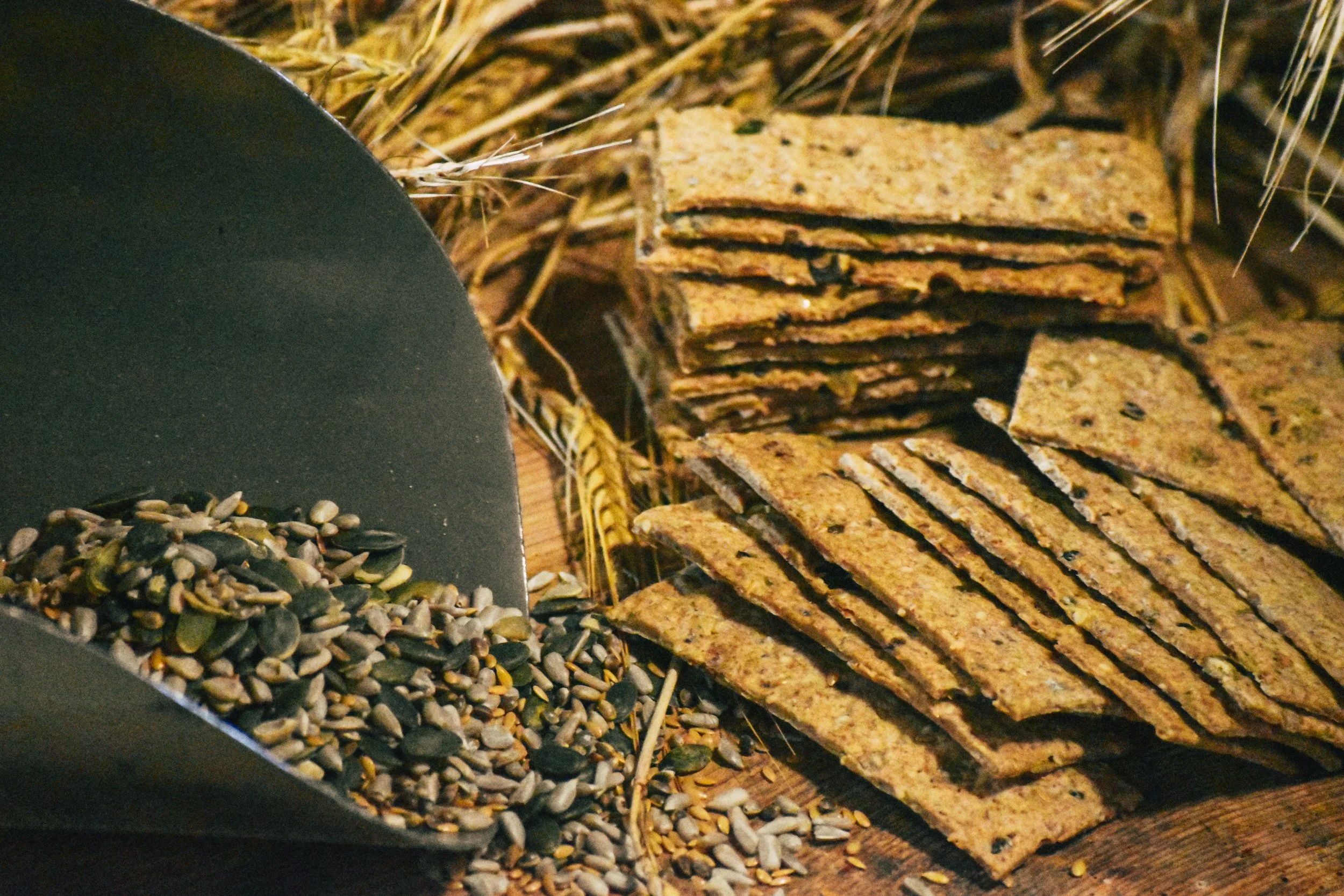 Stacked rectangular seed crackers on a wooden surface with a grain of wheat and mixed seeds in the foreground.