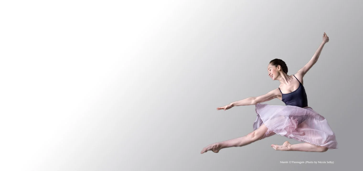 Ballet dancer in mid-air pose wearing a navy leotard and pink tutu, against a gray background.
