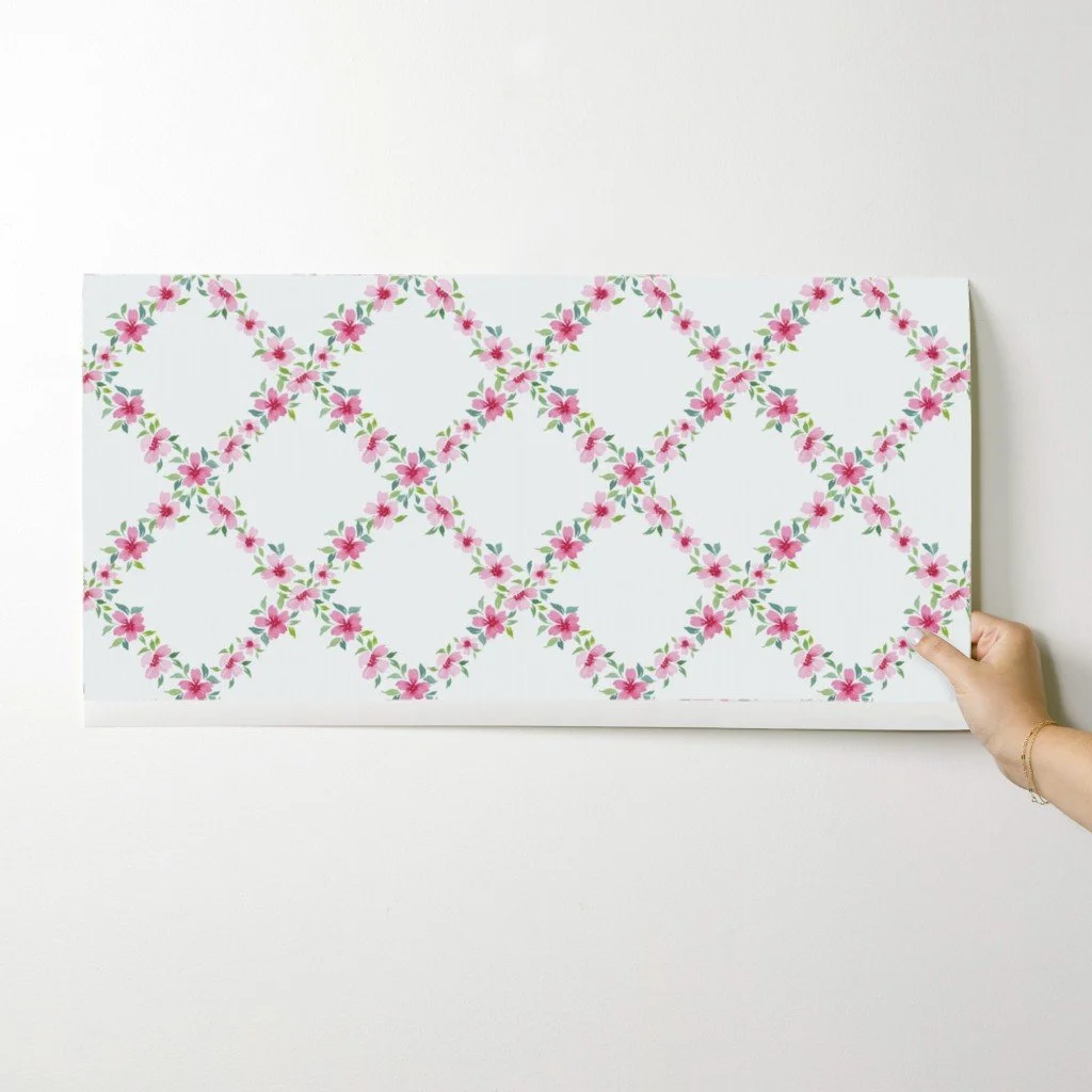 Hand holding a sample strip of wallpaper featuring a repeating diamond lattice pattern made of small pink watercolor flowers with green leaves on a white background.