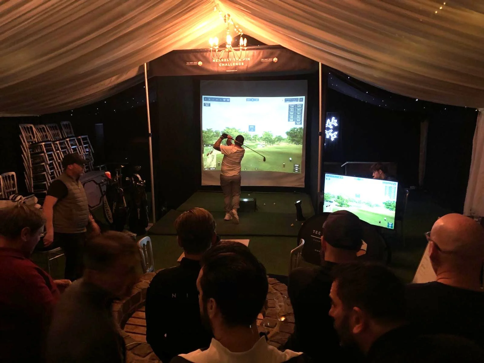 People watching a golf simulator inside a tent, with a person swinging a golf club on a screen, and multiple chairs stacked on the side.