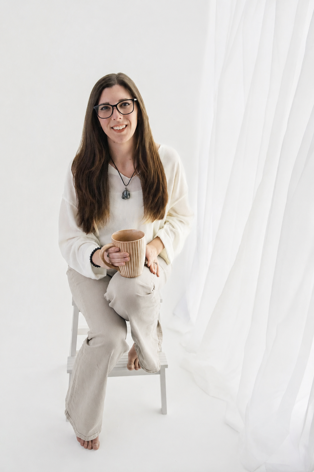 Smiling woman holding a ceramic mug during a natural light personal branding session in a bright white studio with soft curtains, photographed by El & Em Photography in Madison Heights, Michigan.