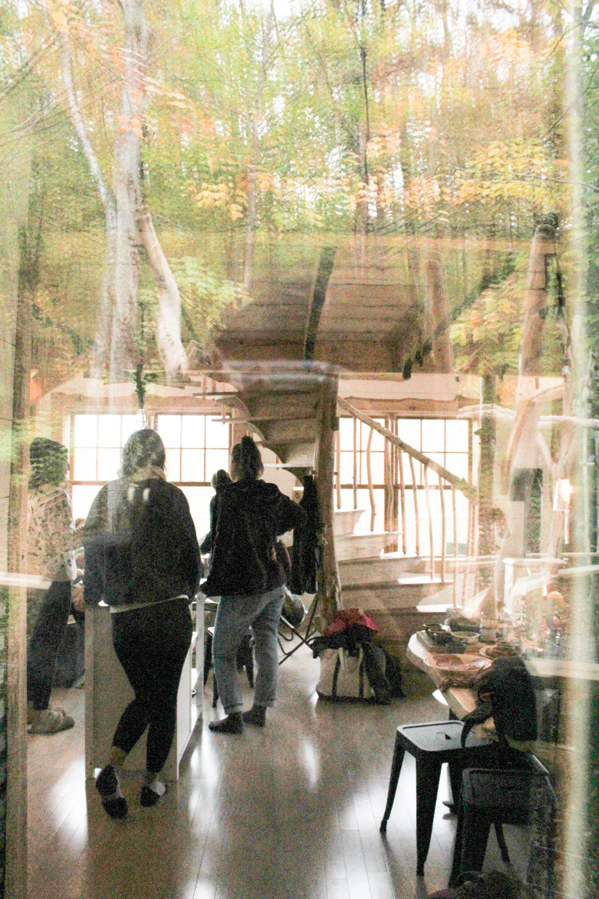 People are standing and talking inside a room with large windows. The room has unfinished wooden stairs and a wall with a mural of trees and nature.